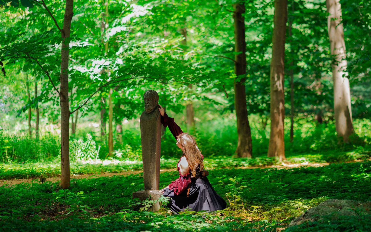 A dramatic pose against a forest backdrop features flowing burgundy and black period costume.