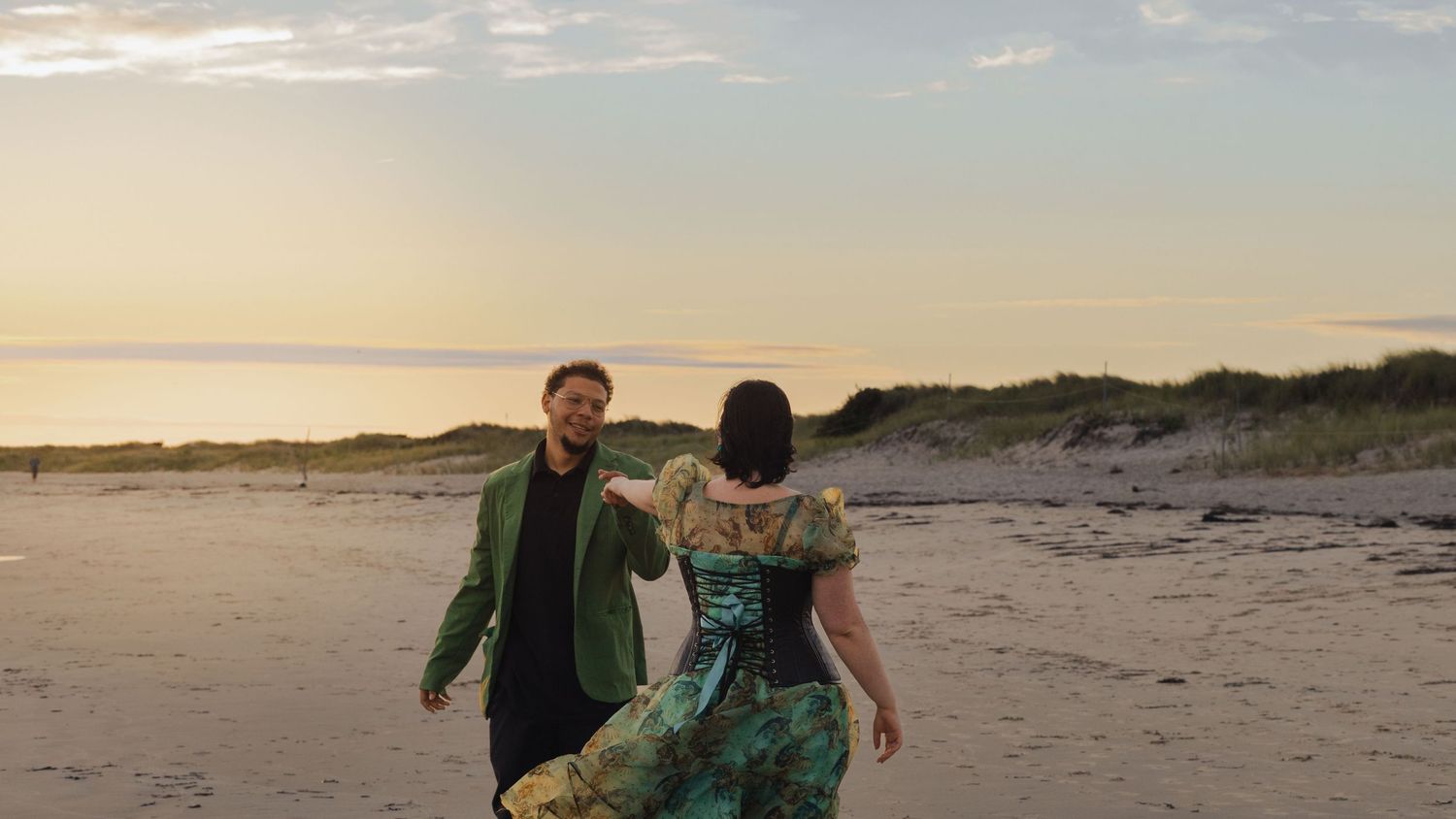A couple strolls together on a sandy beach during golden hour with dunes in the background.