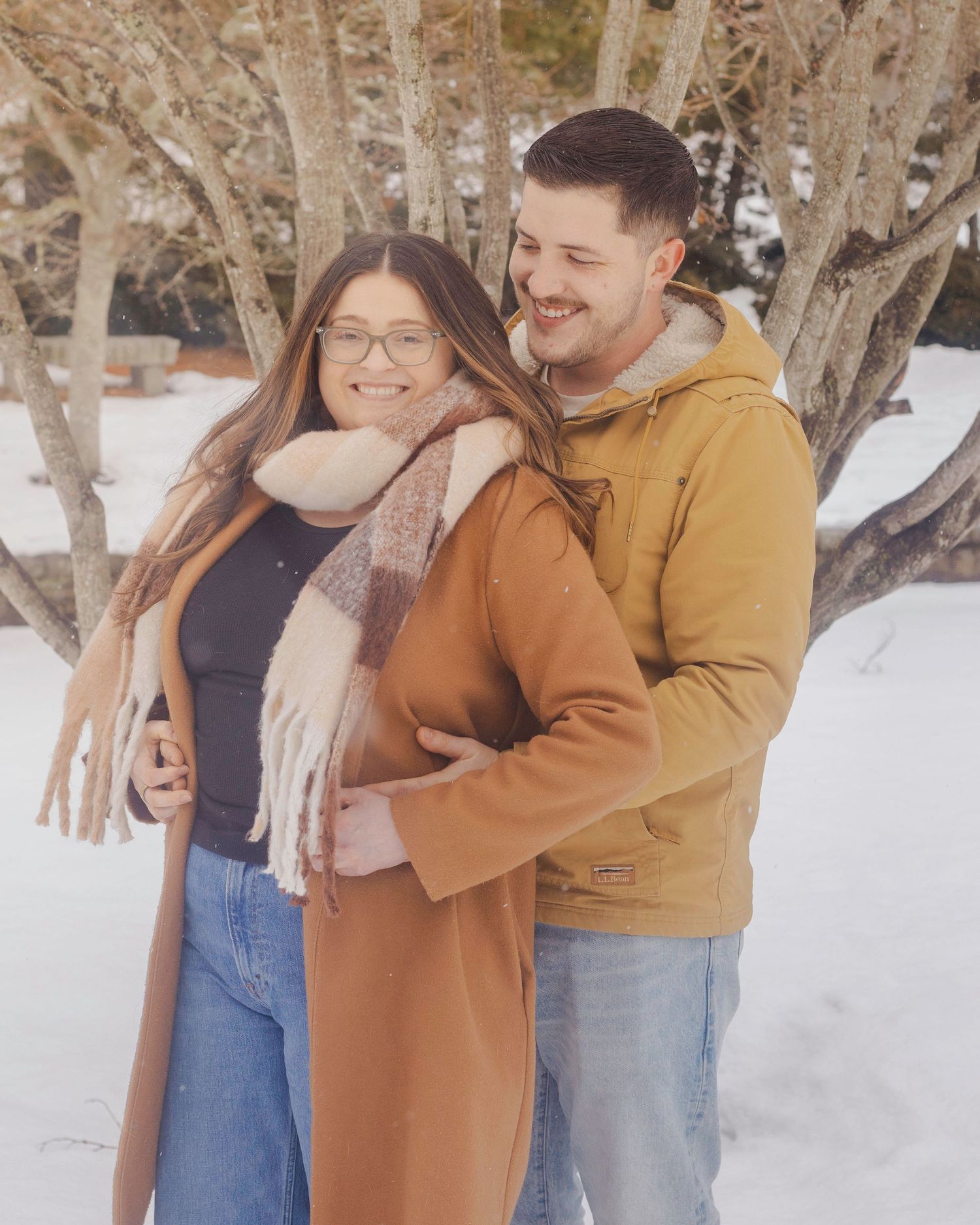Couple smiling and embracing outdoors in winter, woman in brown coat and scarf, man in yellow hoodie, snowy trees behind.