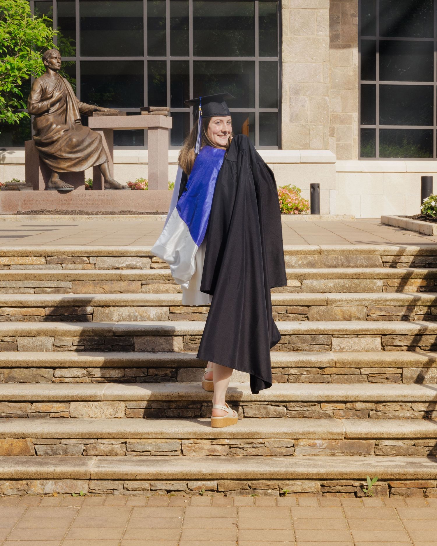 Graduate in black cap and gown with blue sash poses on stone steps outside university building.