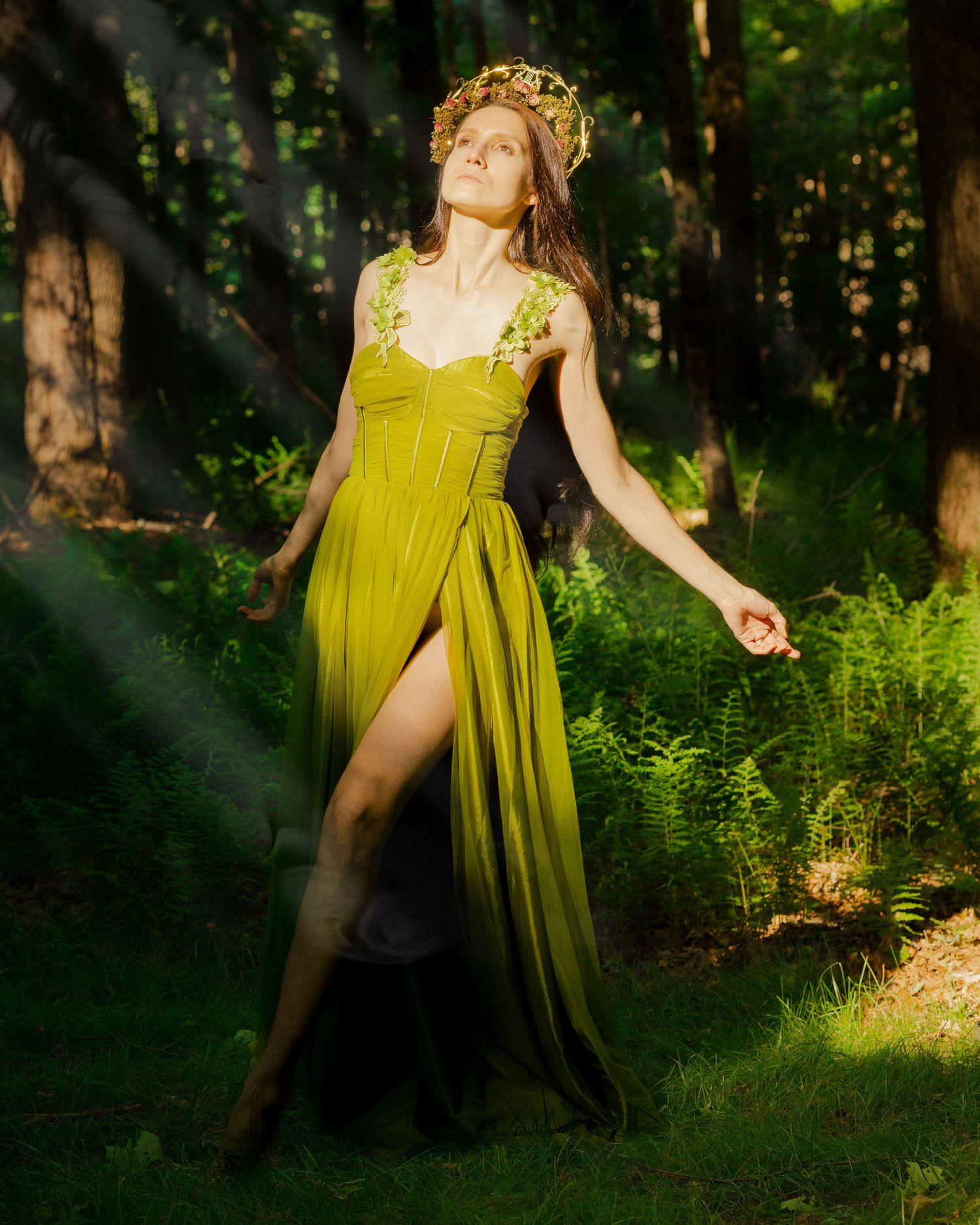 Woman in olive green gown and floral crown poses gracefully in sunlit forest.