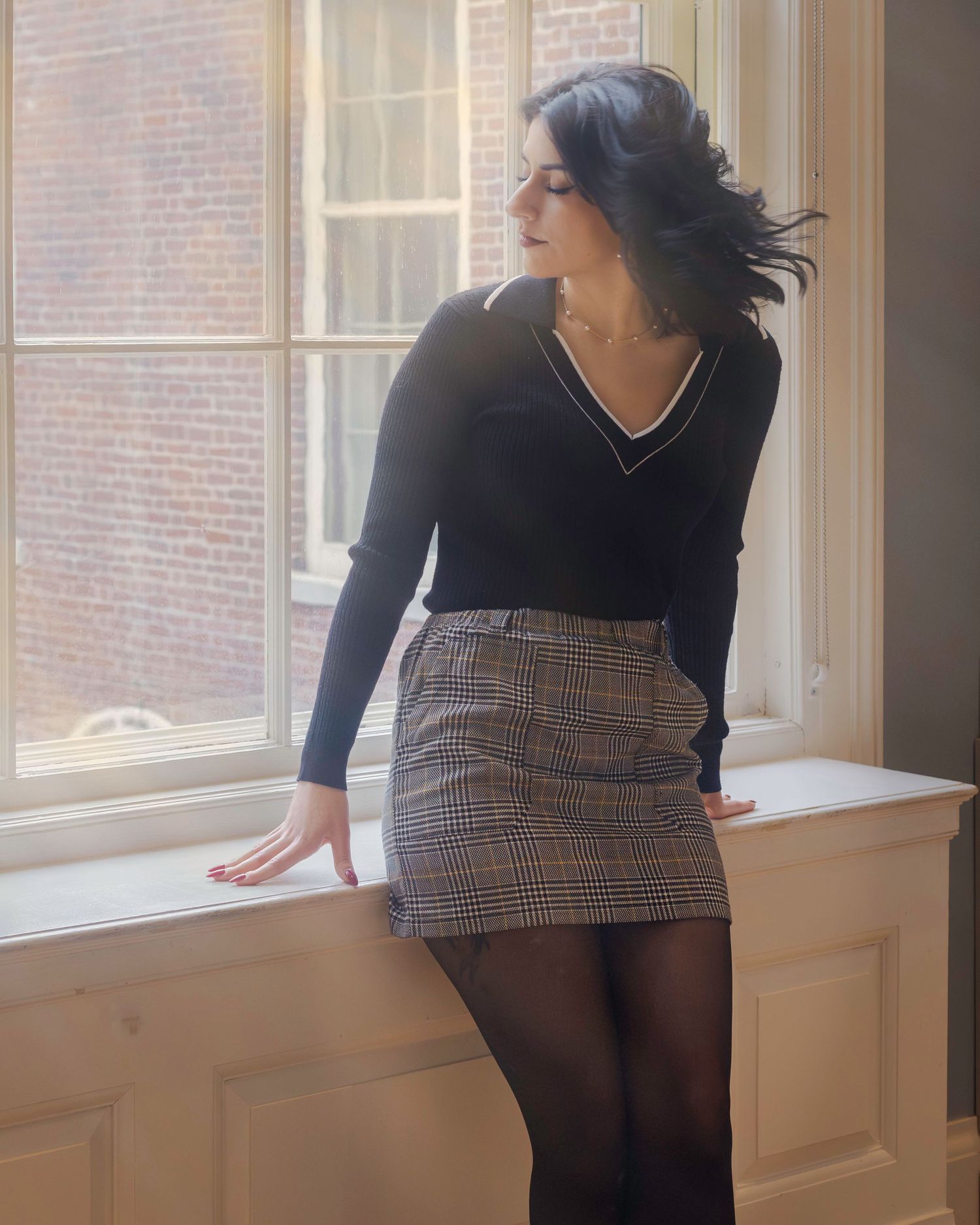 Woman in black V-neck sweater and plaid mini skirt with black tights posing by a large window.