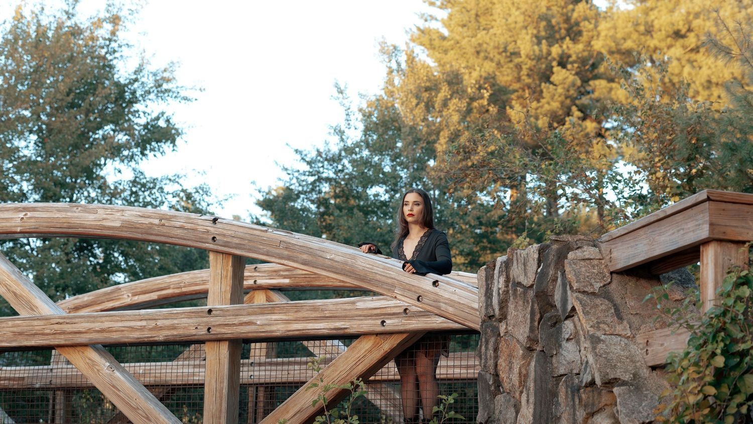 Woman standing on a rustic wooden bridge surrounded by lush trees and stone walls in a scenic outdoor setting.