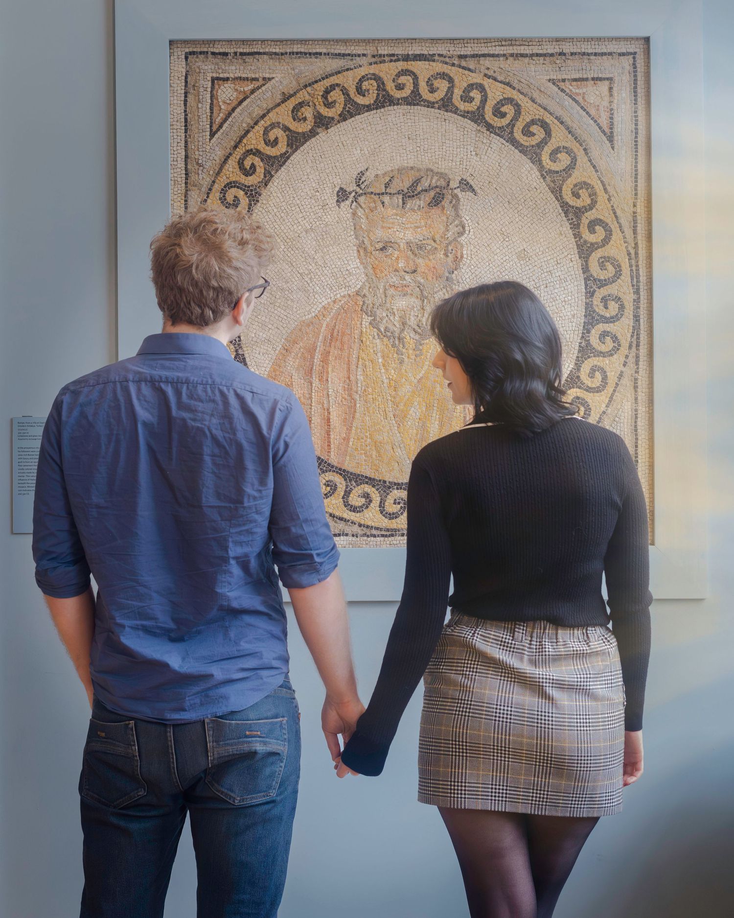 A couple holds hands while viewing an ancient Roman mosaic portrait artwork in a museum gallery.