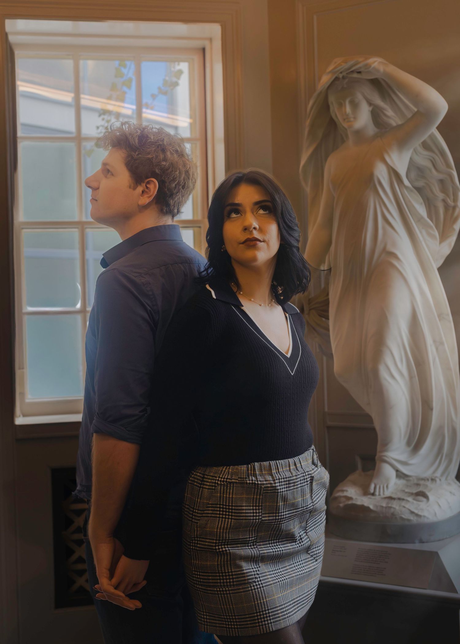 Two people in matching navy V-neck tops and plaid skirts posing near a marble statue indoors.