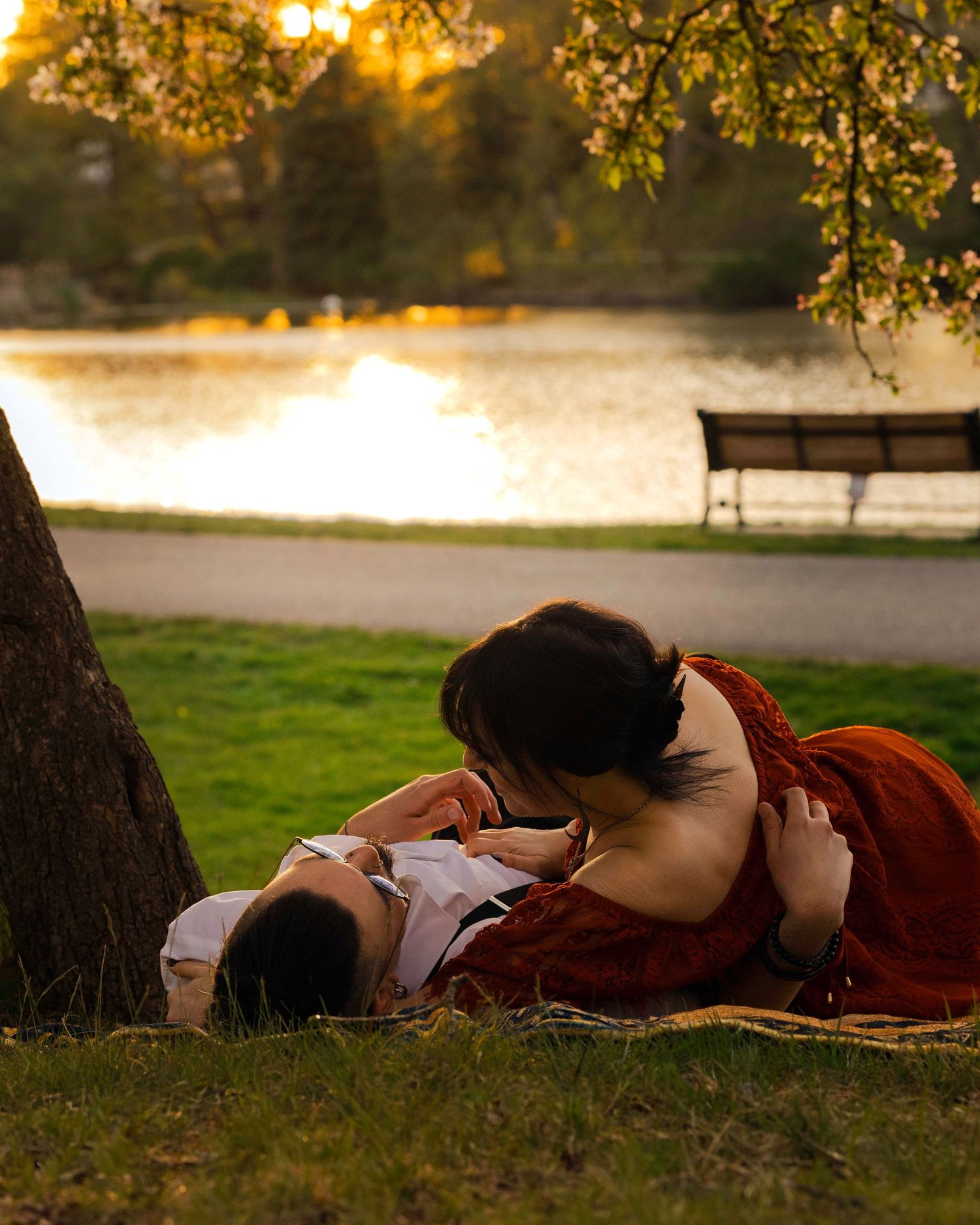 Romantic couple lying on grass by a lake at sunset, sharing an intimate moment near a tree in a park.
