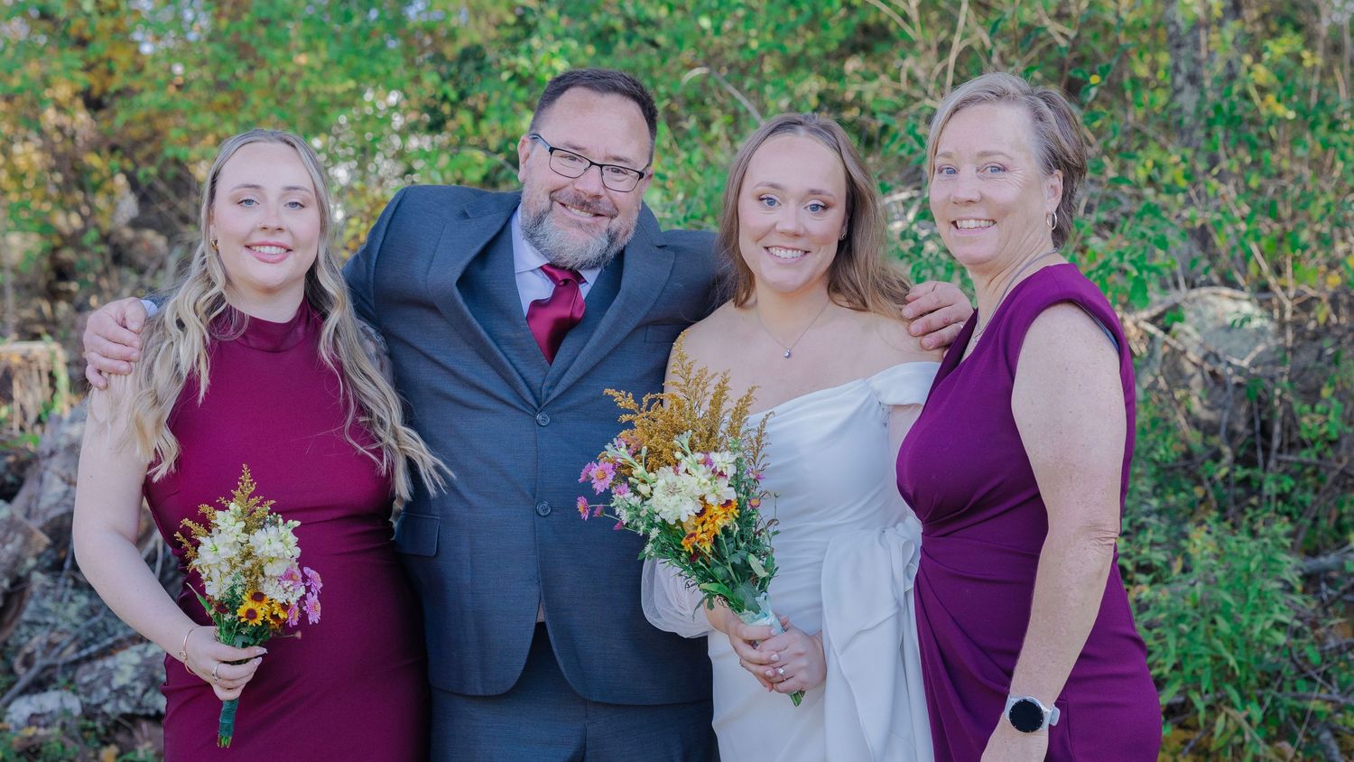 Wedding party poses in purple and gray attire among green foliage.