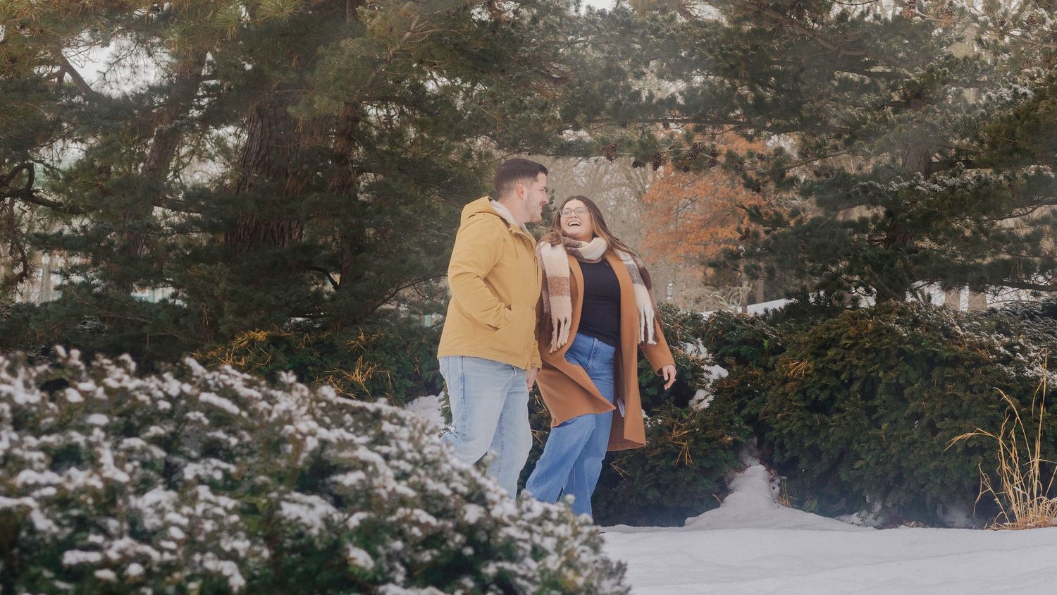 Couple walking together in snowy winter landscape surrounded by evergreen trees on a sunny day.