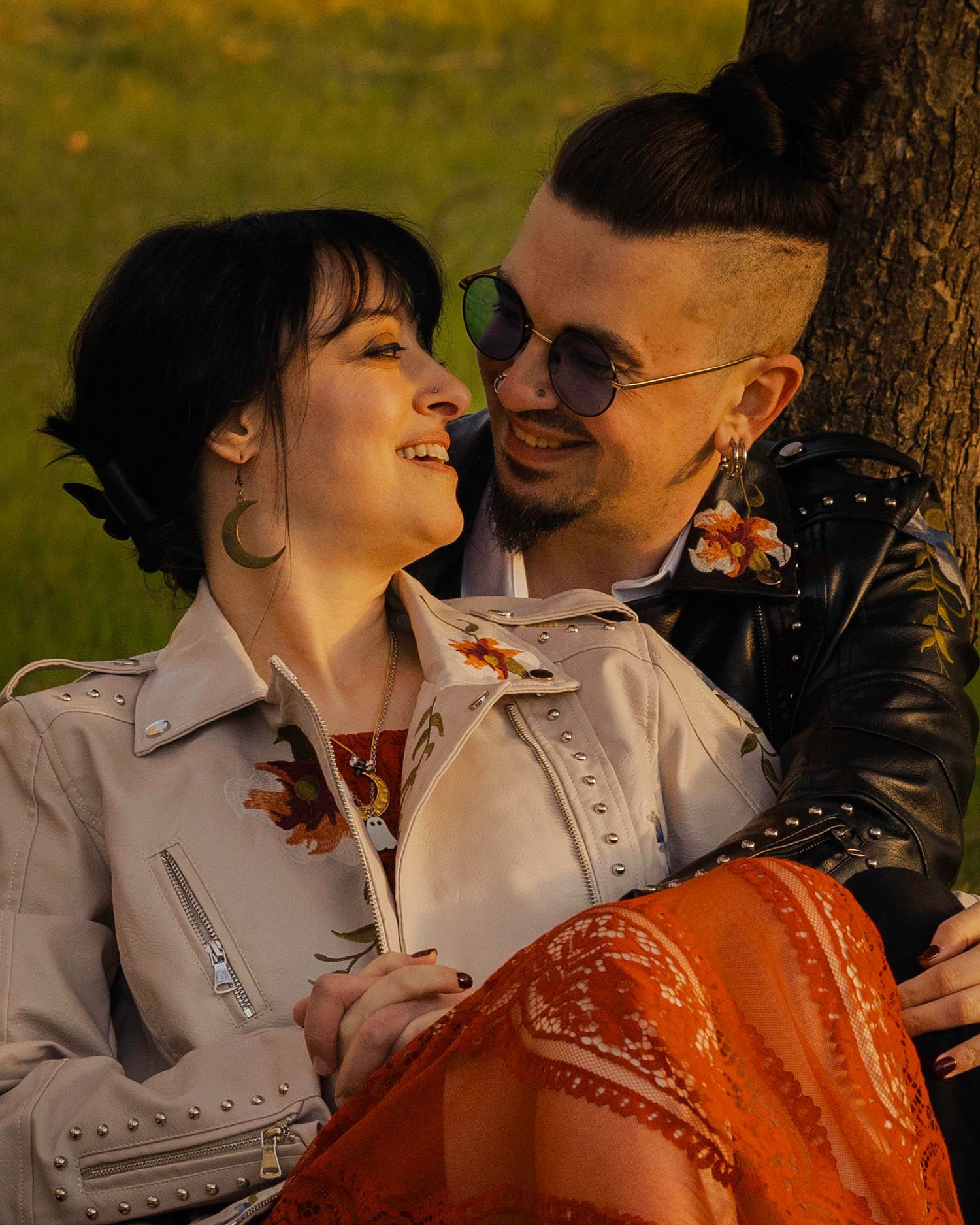 A smiling couple in edgy leather jackets share a romantic moment leaning against a tree outdoors.