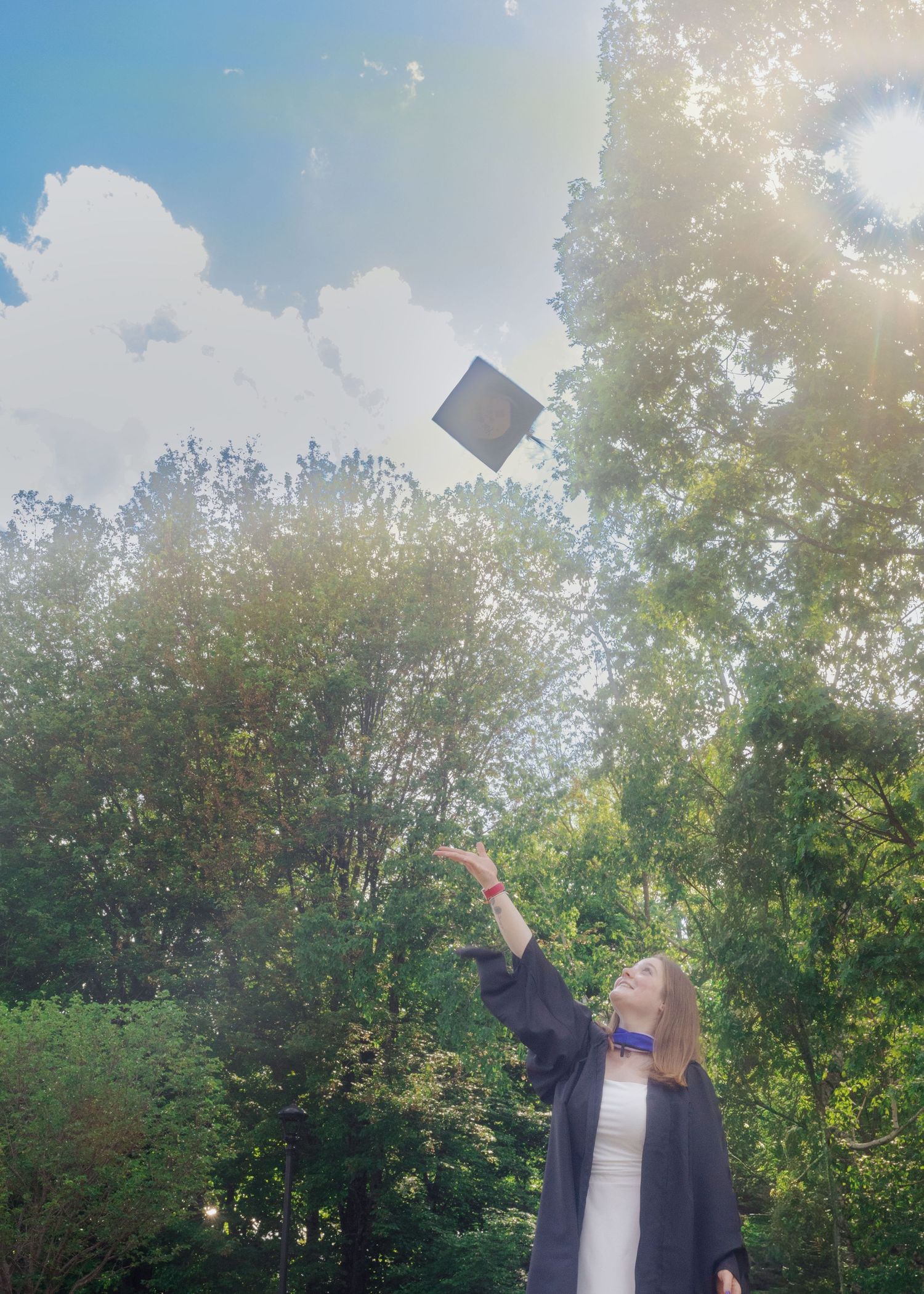 Graduate in cap and gown tossing mortarboard into sunny sky surrounded by lush green trees.