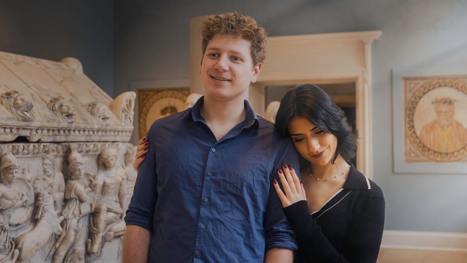A young couple smiling together in a museum with classical sculptures and artwork in the background.