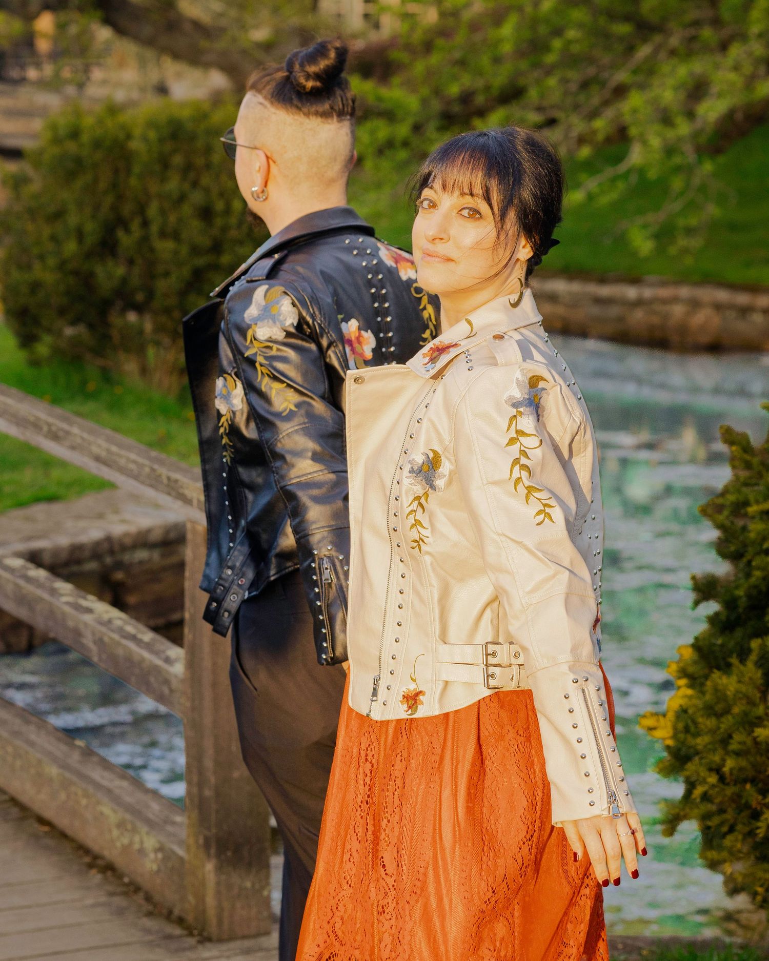 Two people in embroidered leather jackets pose back-to-back outdoors near a pond in a lush green park.