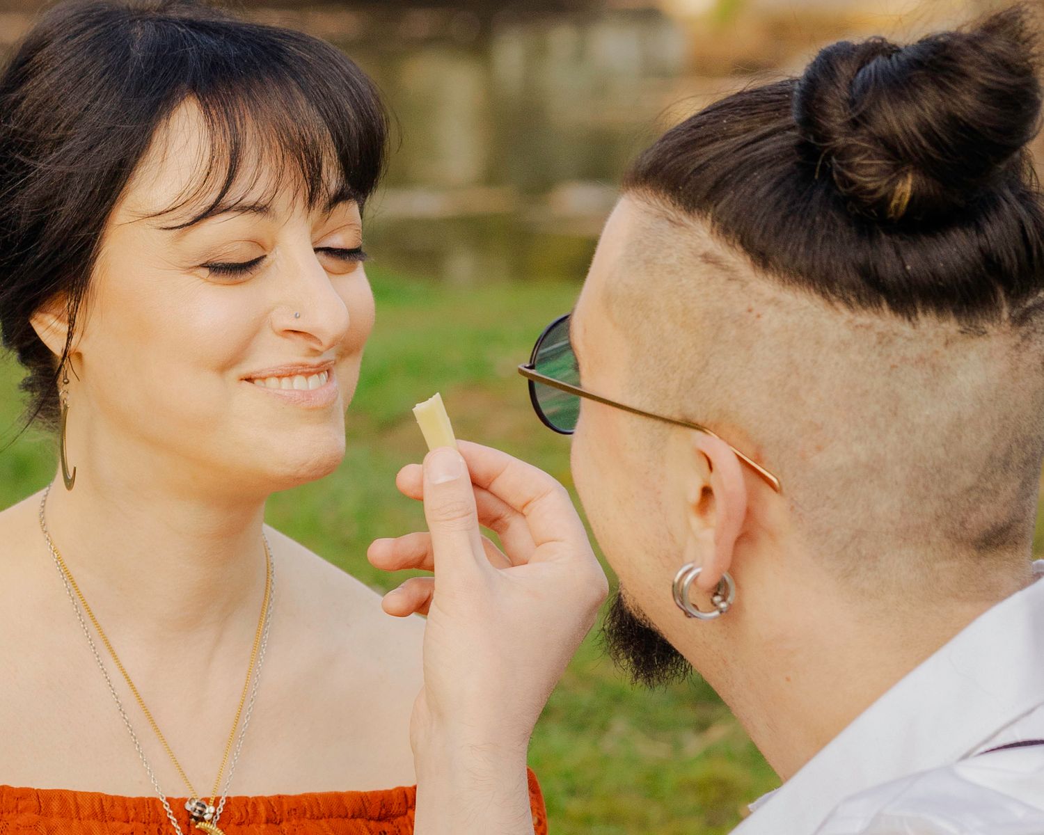 A smiling woman receives a piece of gum from a bearded man with a bun hairstyle and sunglasses outdoors.