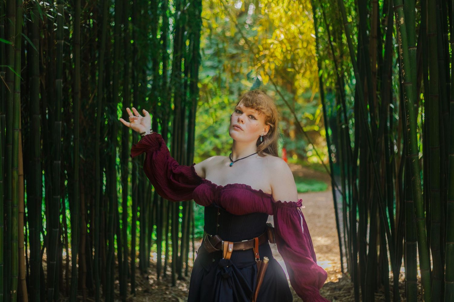 A person in medieval attire reaches out while standing in a bamboo forest path.