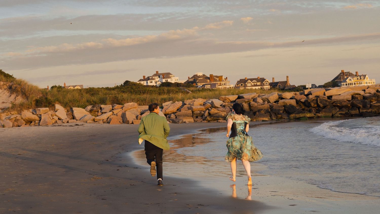 Two figures walk along a scenic beach at sunset with coastal homes visible on the cliffs in the background.
