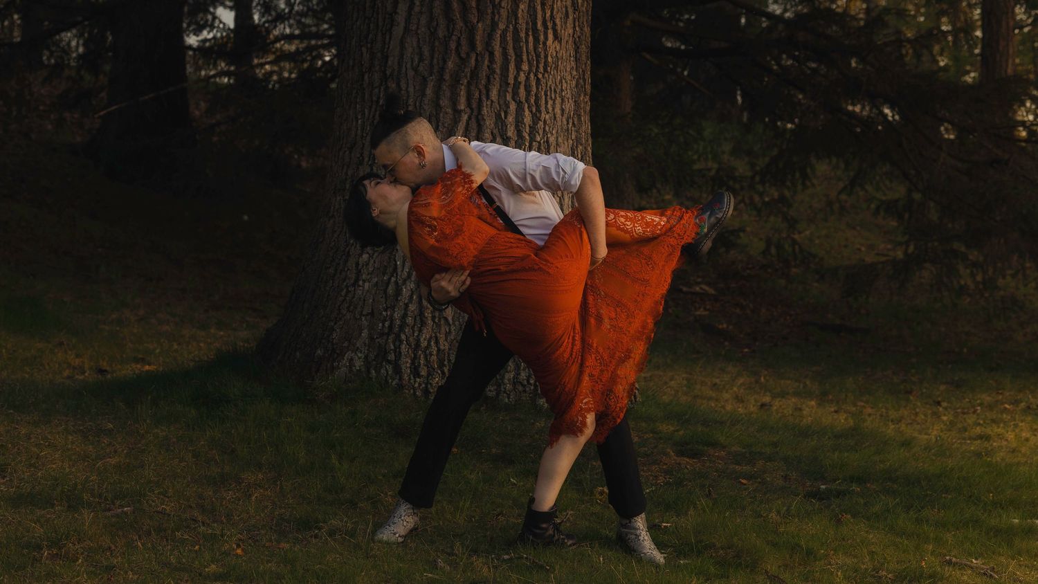 Couple sharing a romantic dip dance pose near a large tree in a sunlit forest, woman in orange dress.