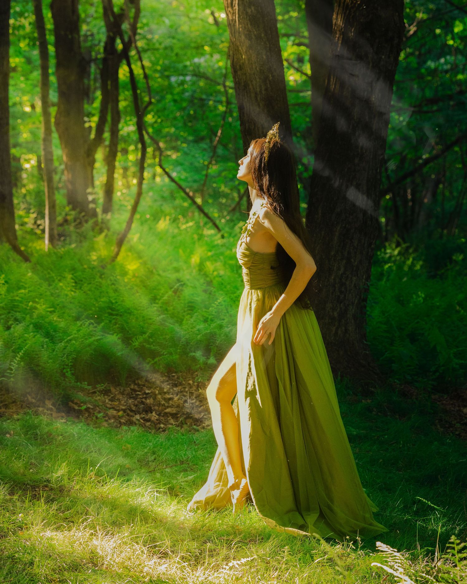 Woman in olive green gown standing in sunlit forest among ferns.
