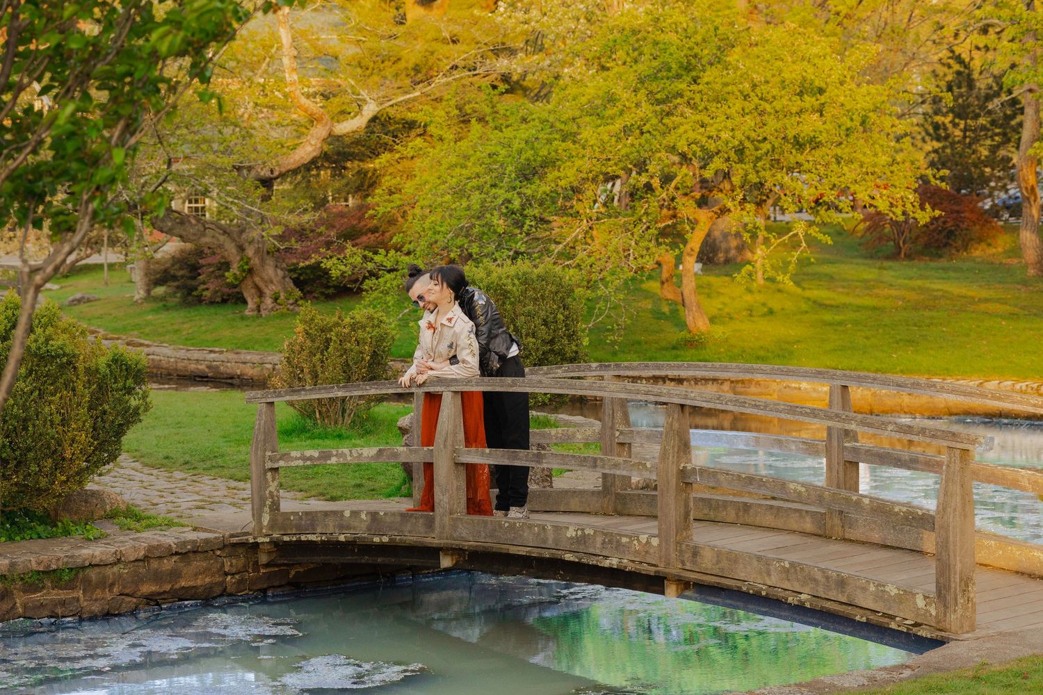 Woman in traditional dress stands on wooden bridge over stream in lush autumn garden with colorful foliage.