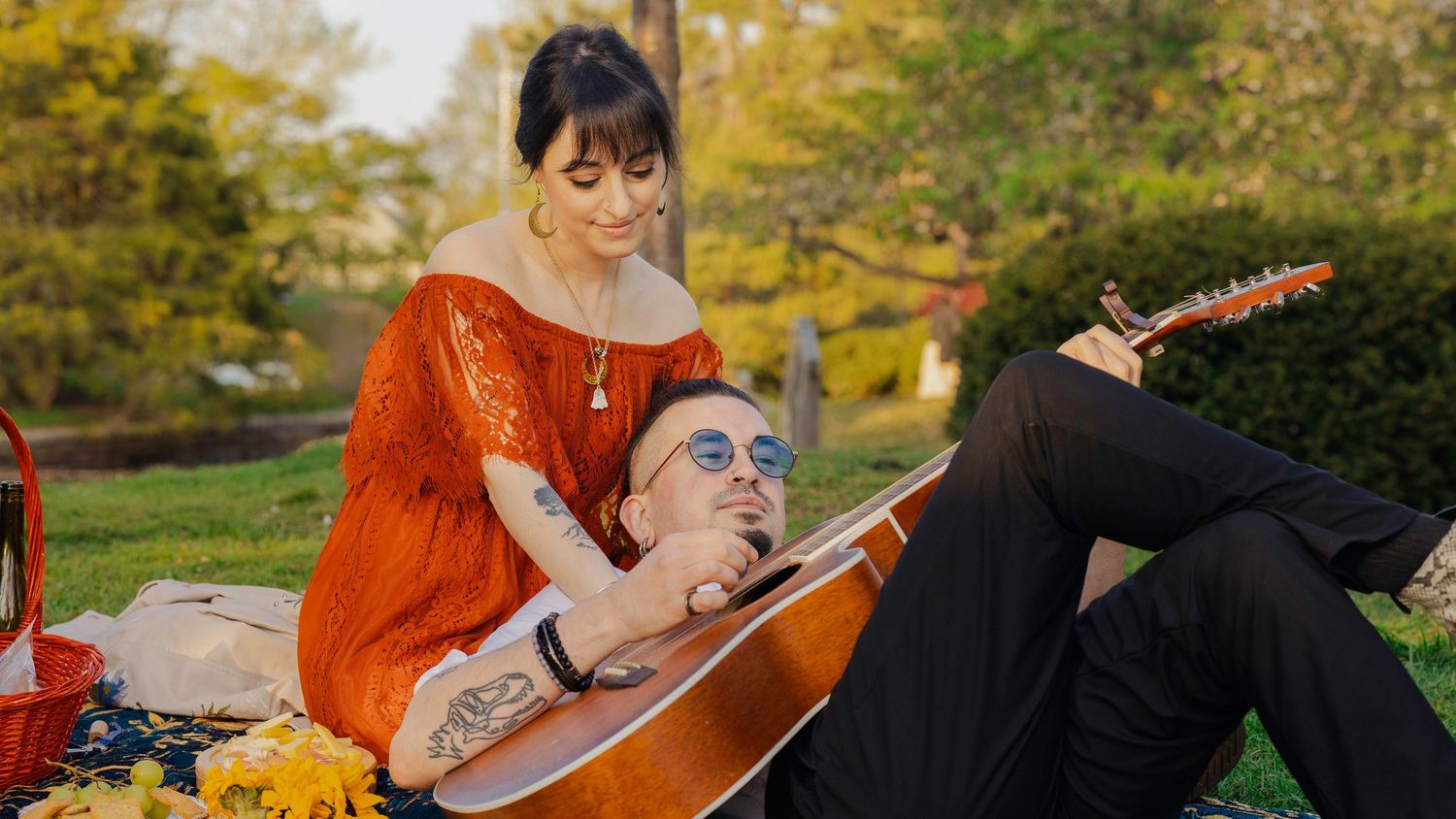 Couple enjoying a romantic picnic outdoors, woman in red dress embracing man playing acoustic guitar.