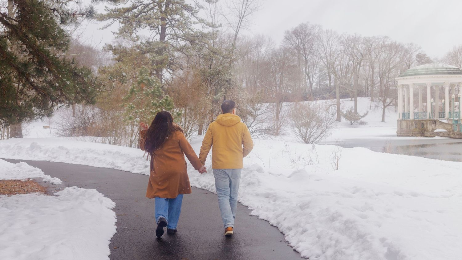 Couple holding hands walking on a snowy park path in winter, wearing colorful coats near a gazebo.