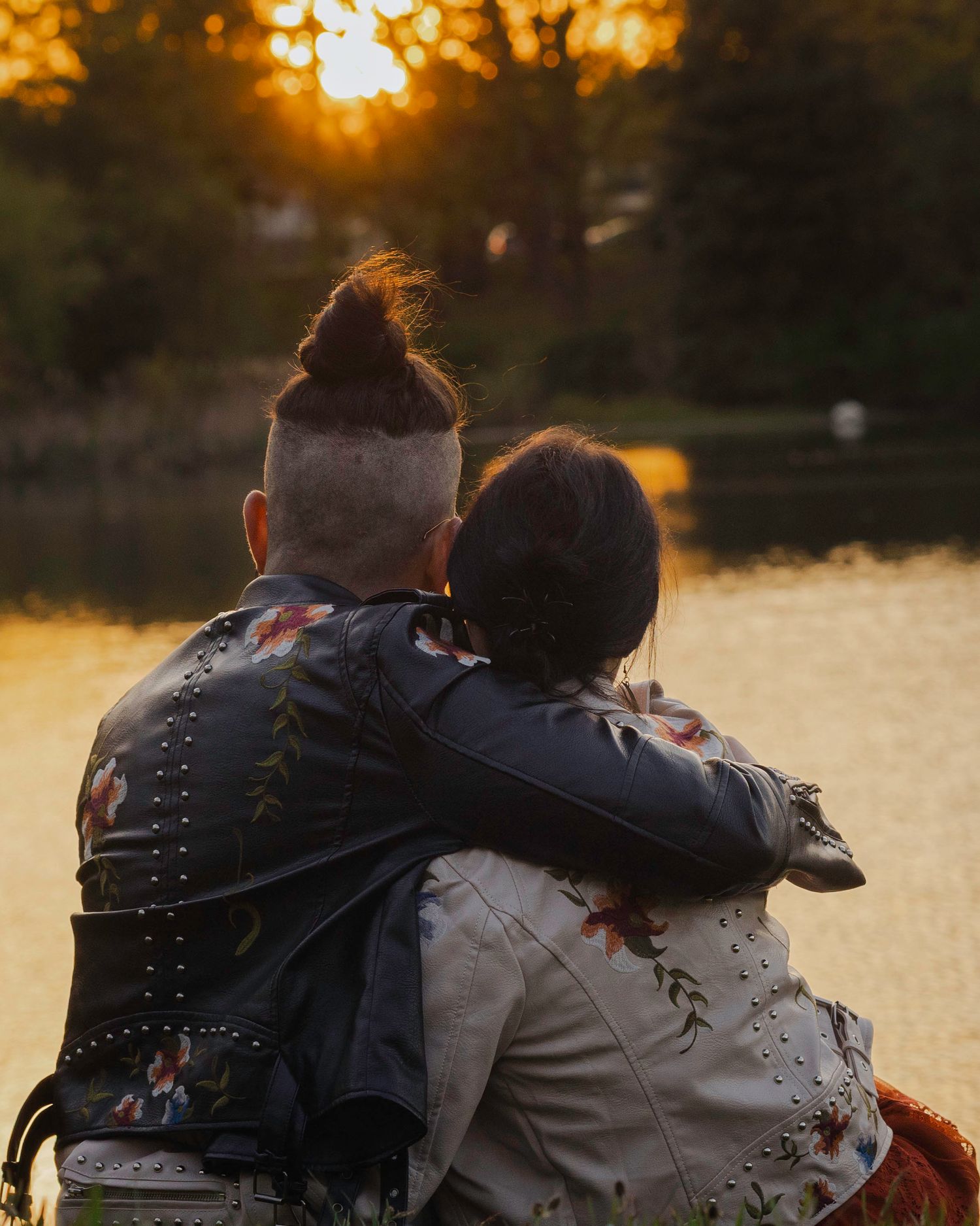Couple embracing at sunset by a lake, wearing matching floral leather jackets, man with undercut hairstyle.