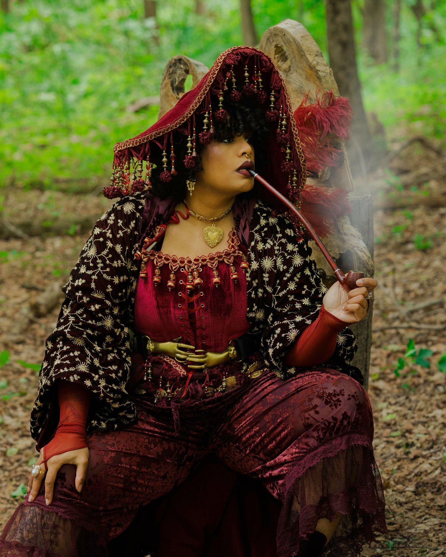 Woman in ornate red and gold gypsy costume smoking a pipe in a forest setting.