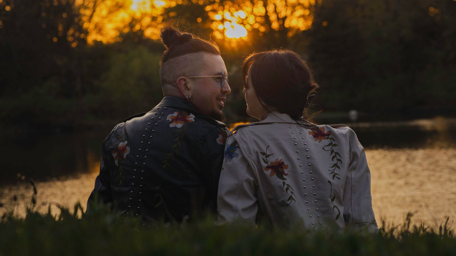 Couple sitting together by a lake at golden sunset, wearing embroidered jackets, sharing a romantic moment outdoors.