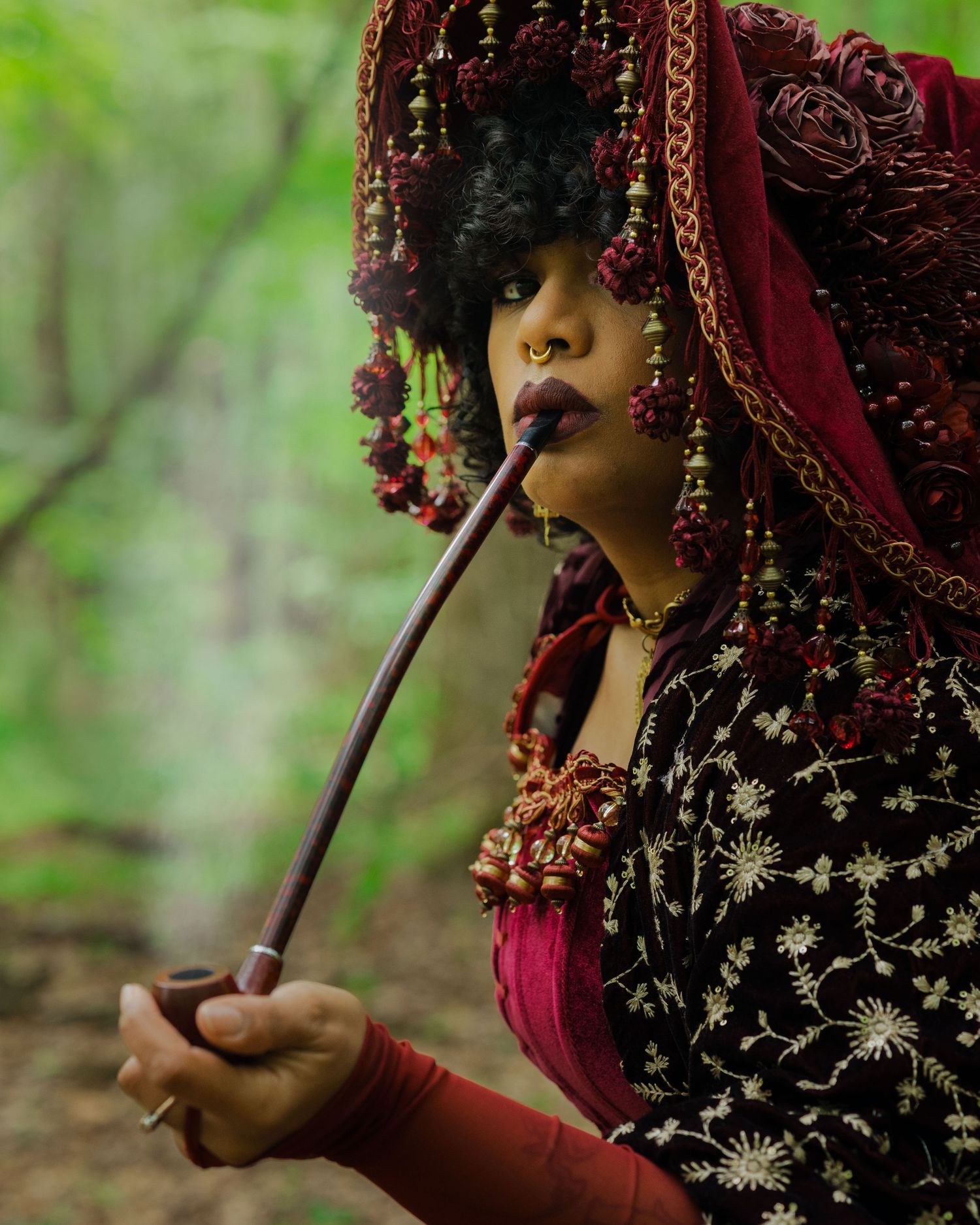 Woman in ornate burgundy headdress and embroidered attire smoking a long pipe in a forest setting.