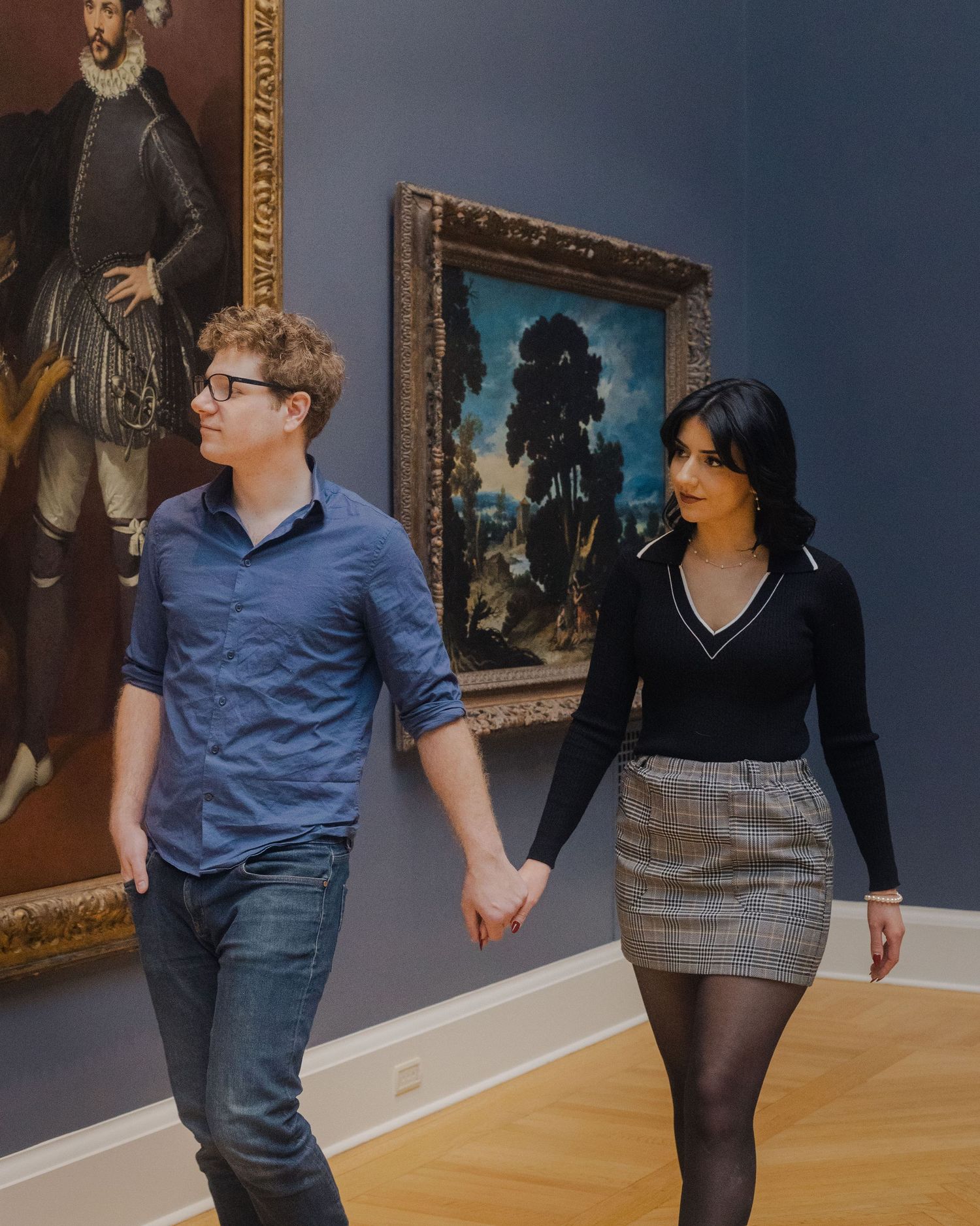 A young couple holds hands while walking through an art museum gallery past classic oil paintings.