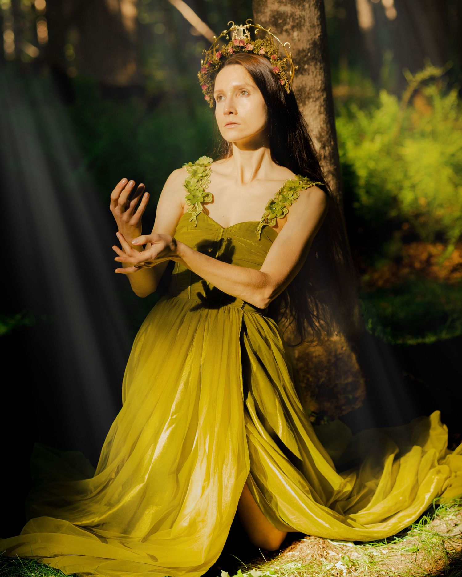 Woman in golden yellow gown and floral crown poses gracefully in a mystical forest setting.