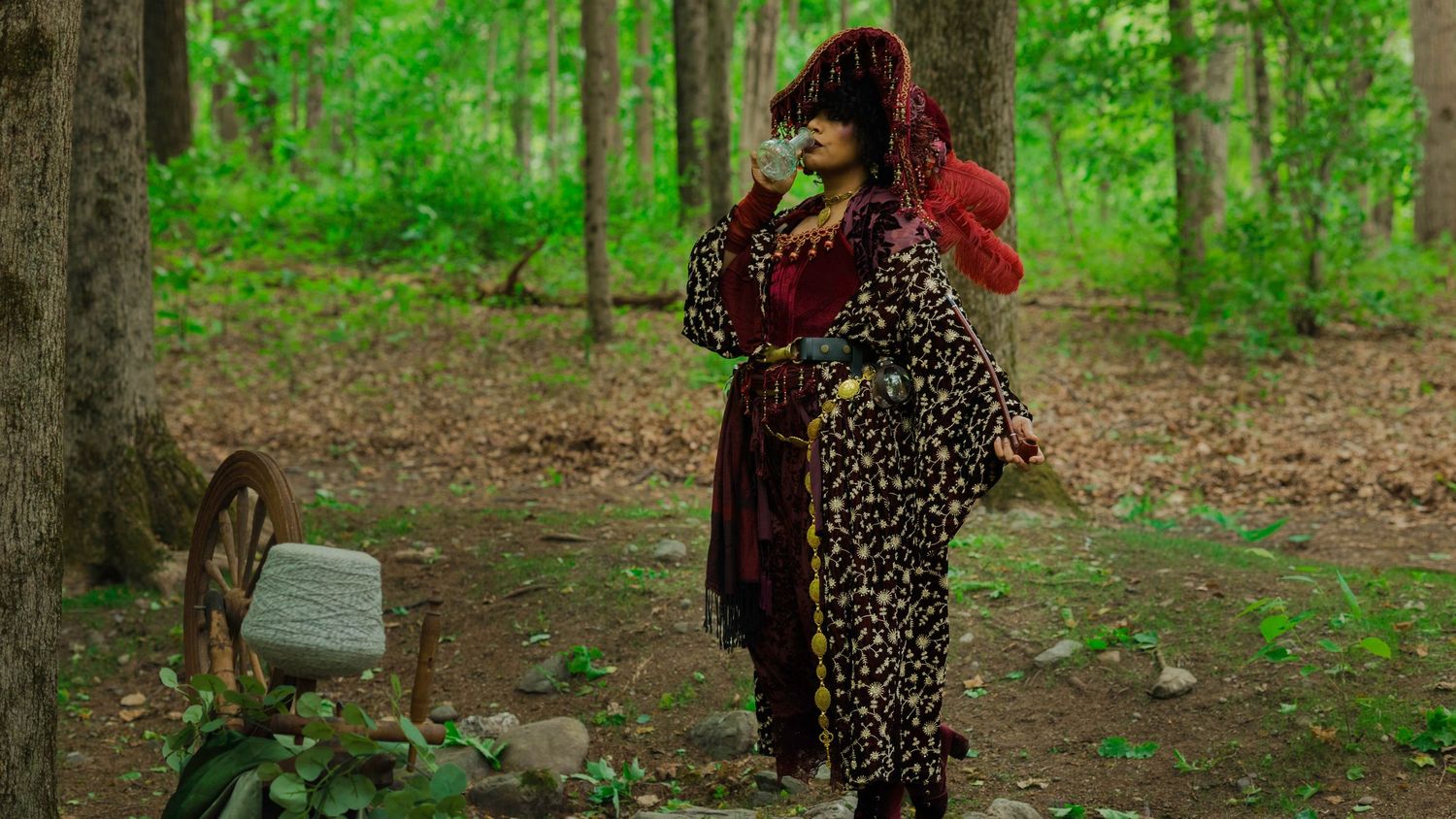 Person in leopard print costume and red hat stands in a forest clearing among trees and rocks.
