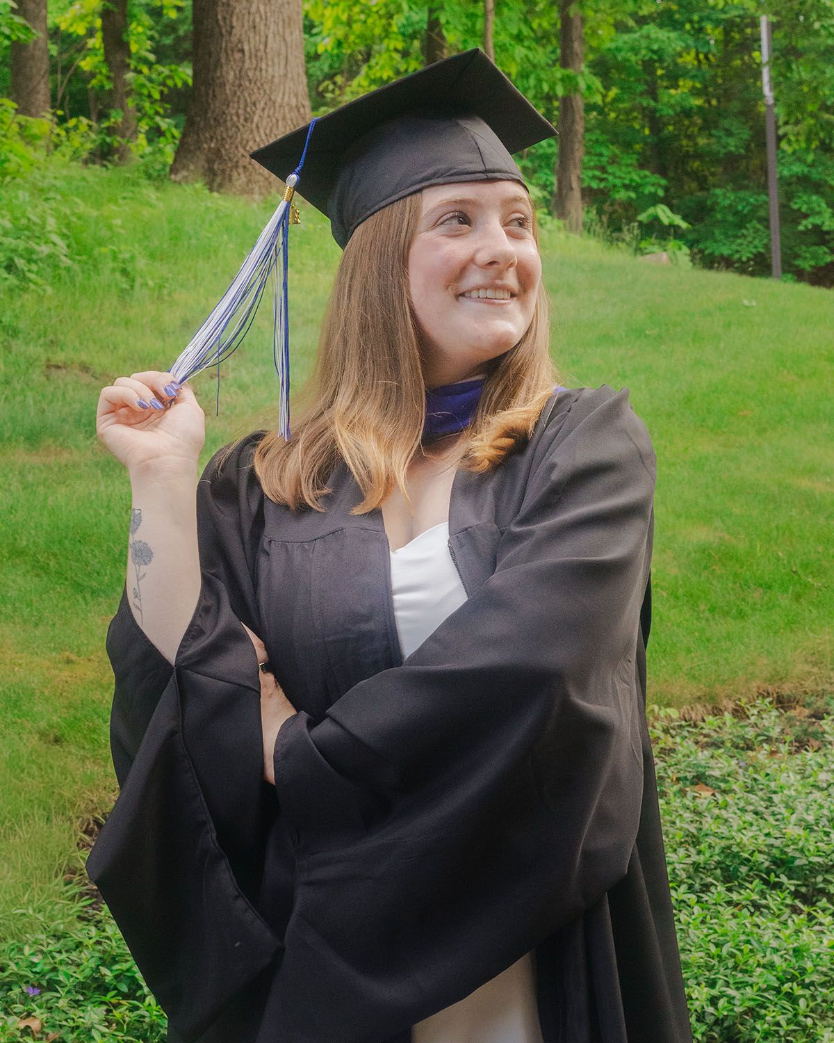 Smiling young woman in black graduation cap and gown holding her tassel in a green outdoor setting.