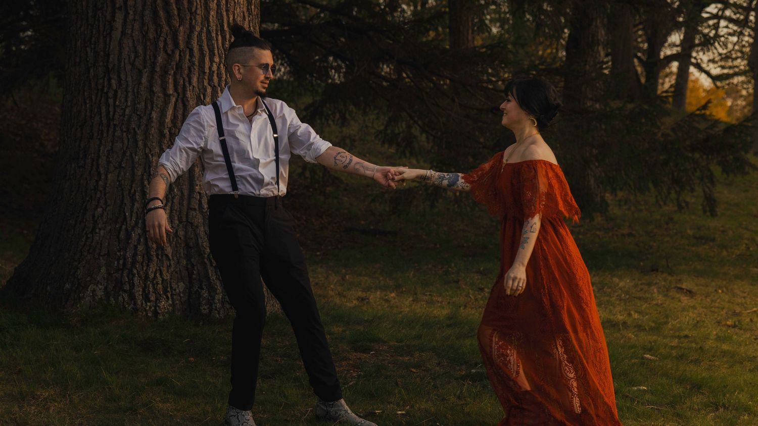 Couple dancing near a tree in a park, woman in orange dress holding hands with man in suspenders at sunset.