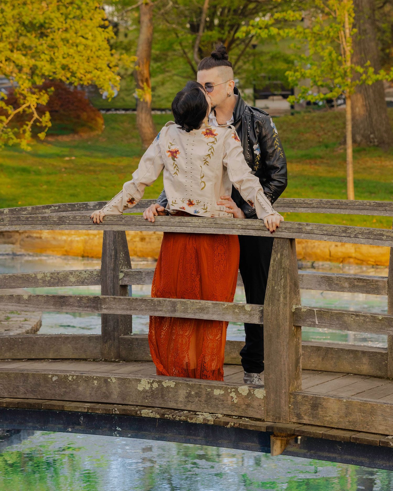 Couple sharing a romantic kiss on a wooden bridge over a pond in a lush green park setting.
