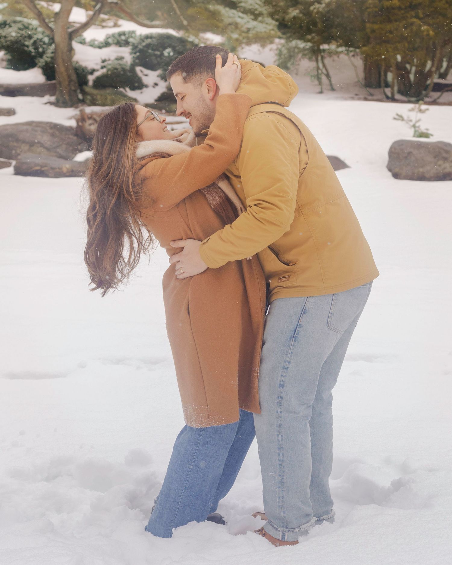 Couple embracing and kissing in snowy winter landscape, wearing warm camel and yellow coats with jeans.