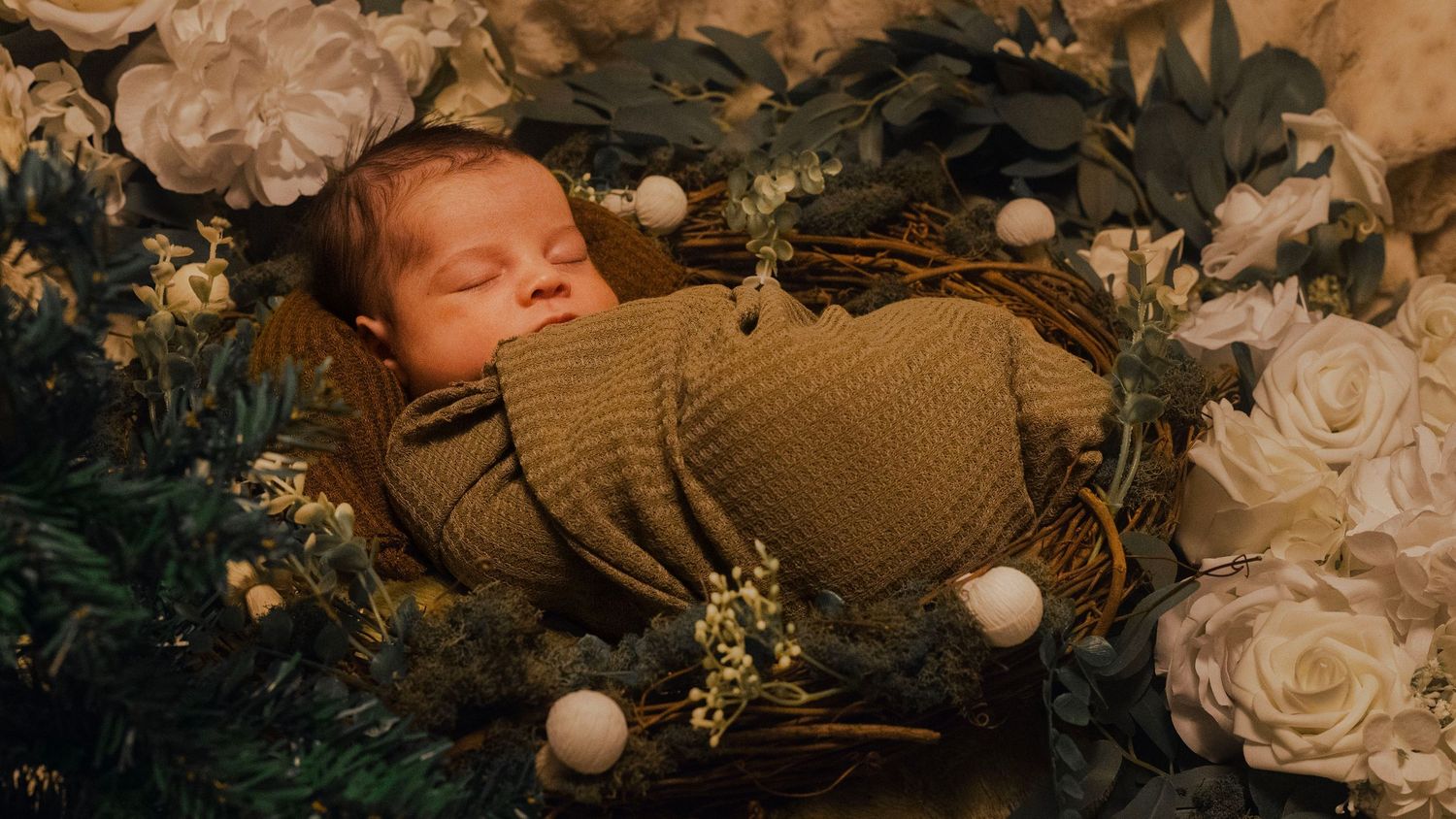 A newborn baby sleeps peacefully surrounded by white roses and soft brown knit blankets in a cozy setting.