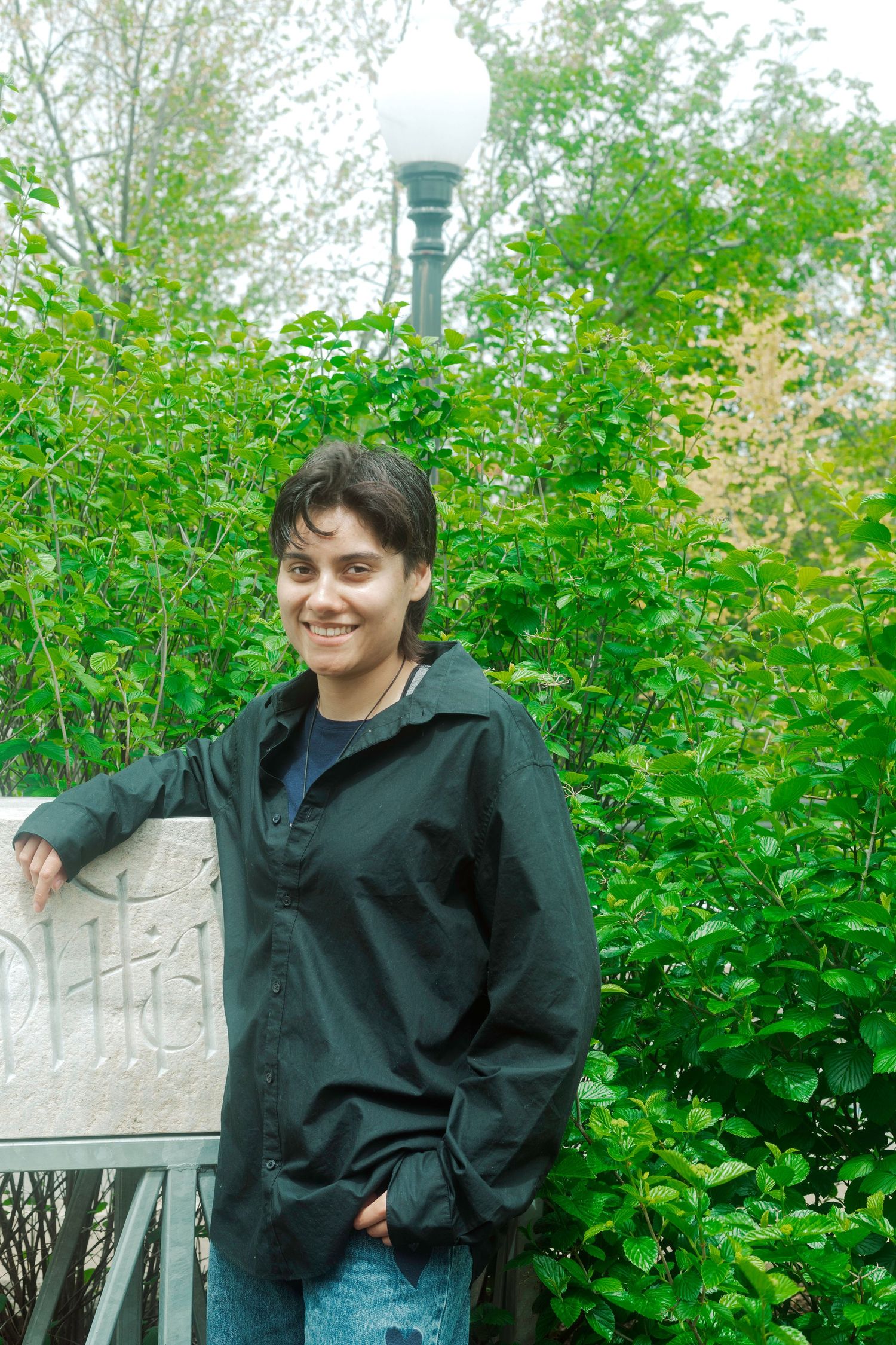 Person in dark jacket poses against lush green garden backdrop with stone wall.