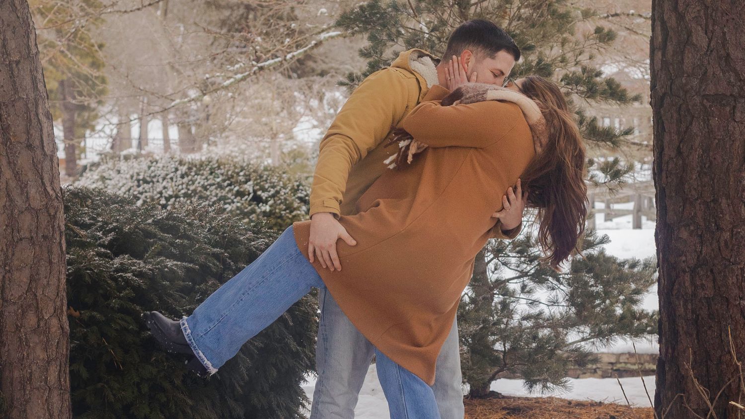 Couple sharing a romantic kiss outdoors in a snowy winter park, man lifting woman in a warm embrace.