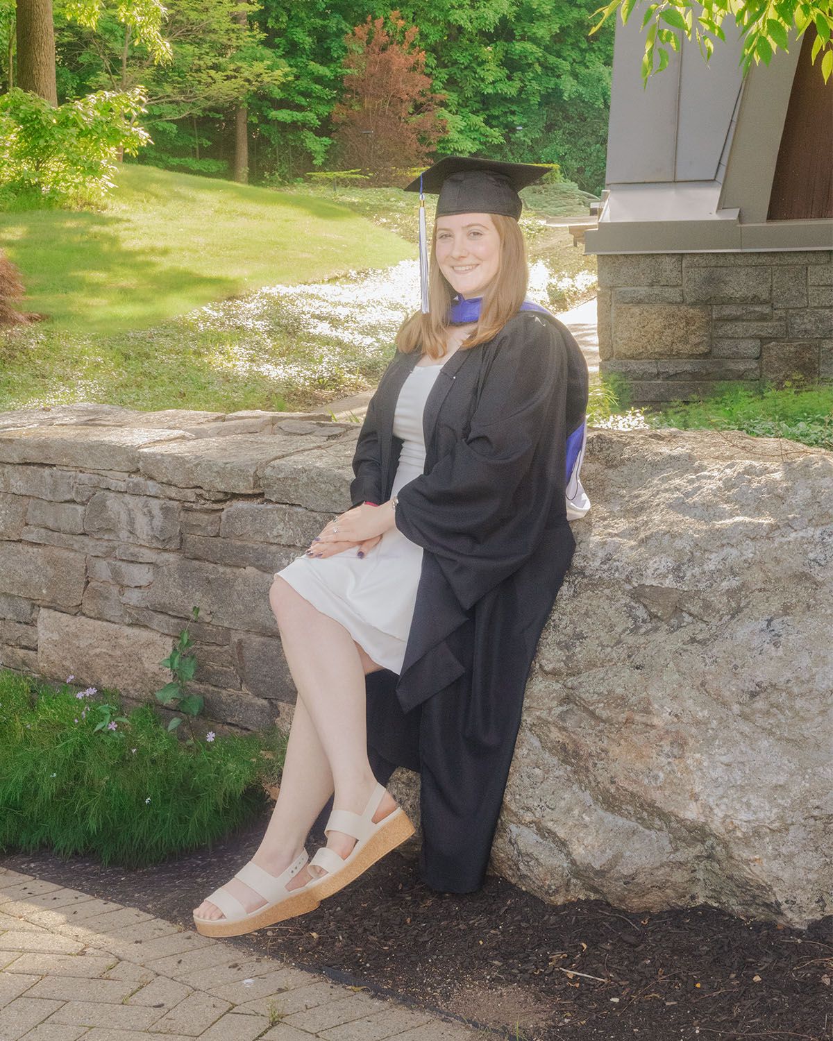 Young woman in black graduation gown and cap sitting on stone wall outdoors, smiling on her graduation day.