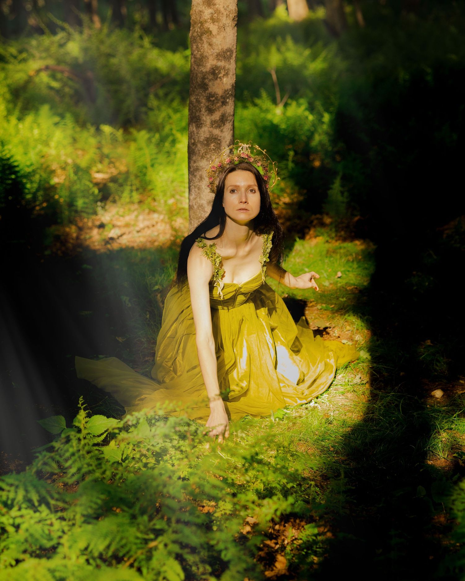 Woman in yellow-green dress and floral crown sits among ferns in enchanted forest.