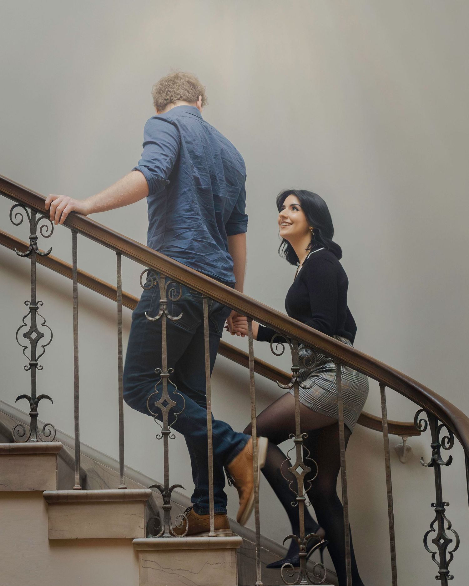 A couple walks up an elegant curved staircase with ornate iron railings in a grand building.