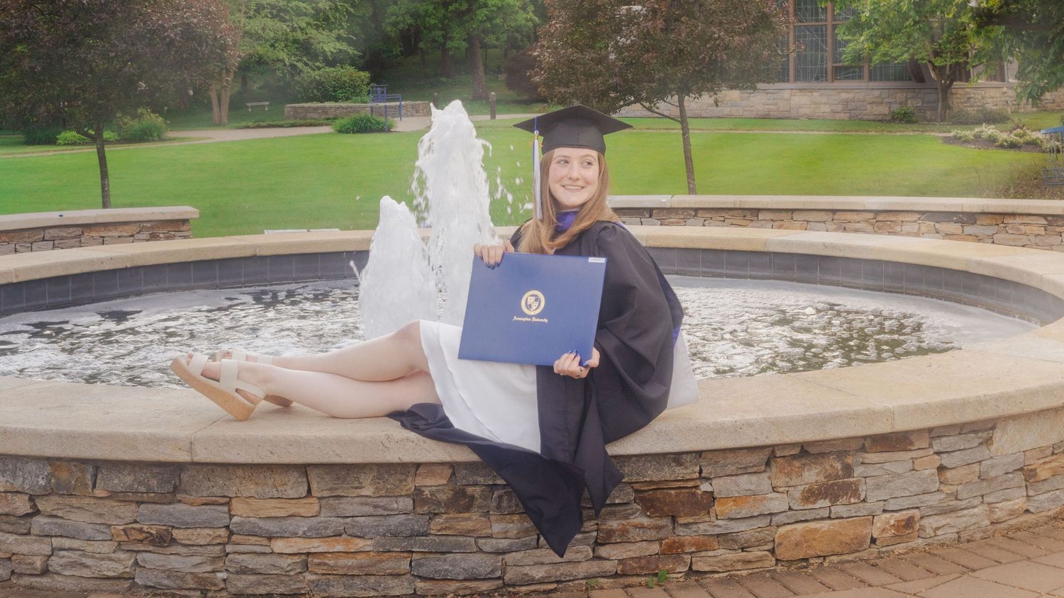 A smiling graduate in cap and gown sits by a stone fountain holding a diploma outdoors.