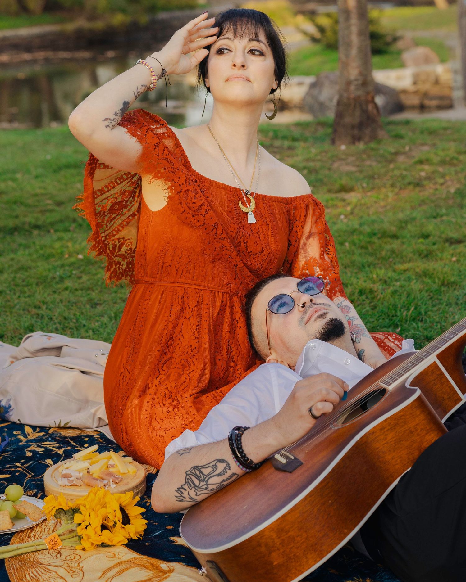 Couple relaxing outdoors on blanket, woman in orange lace dress, man with guitar and sunglasses, picnic setting.