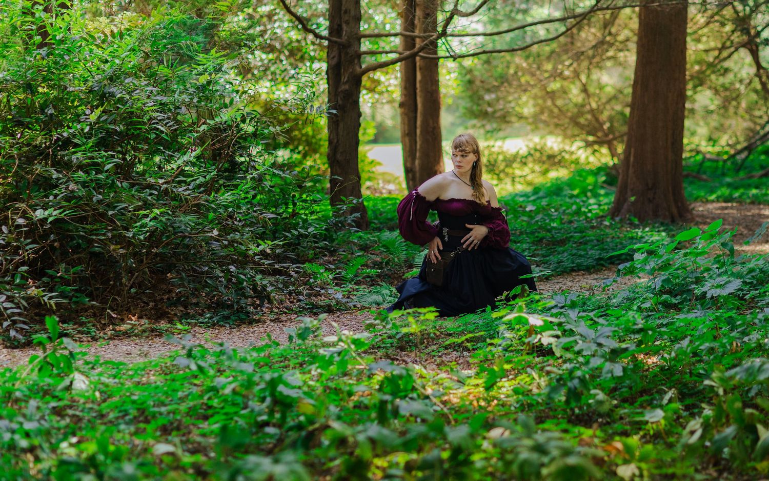 A figure in dark medieval attire crouches in a verdant forest setting.