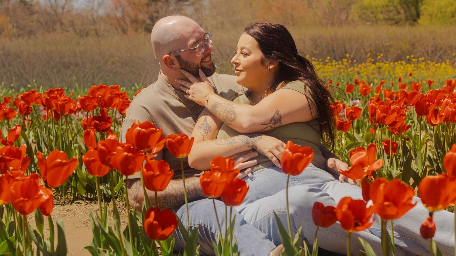 A couple sits romantically among vibrant red tulips in a sunny flower field, sharing a tender moment outdoors.