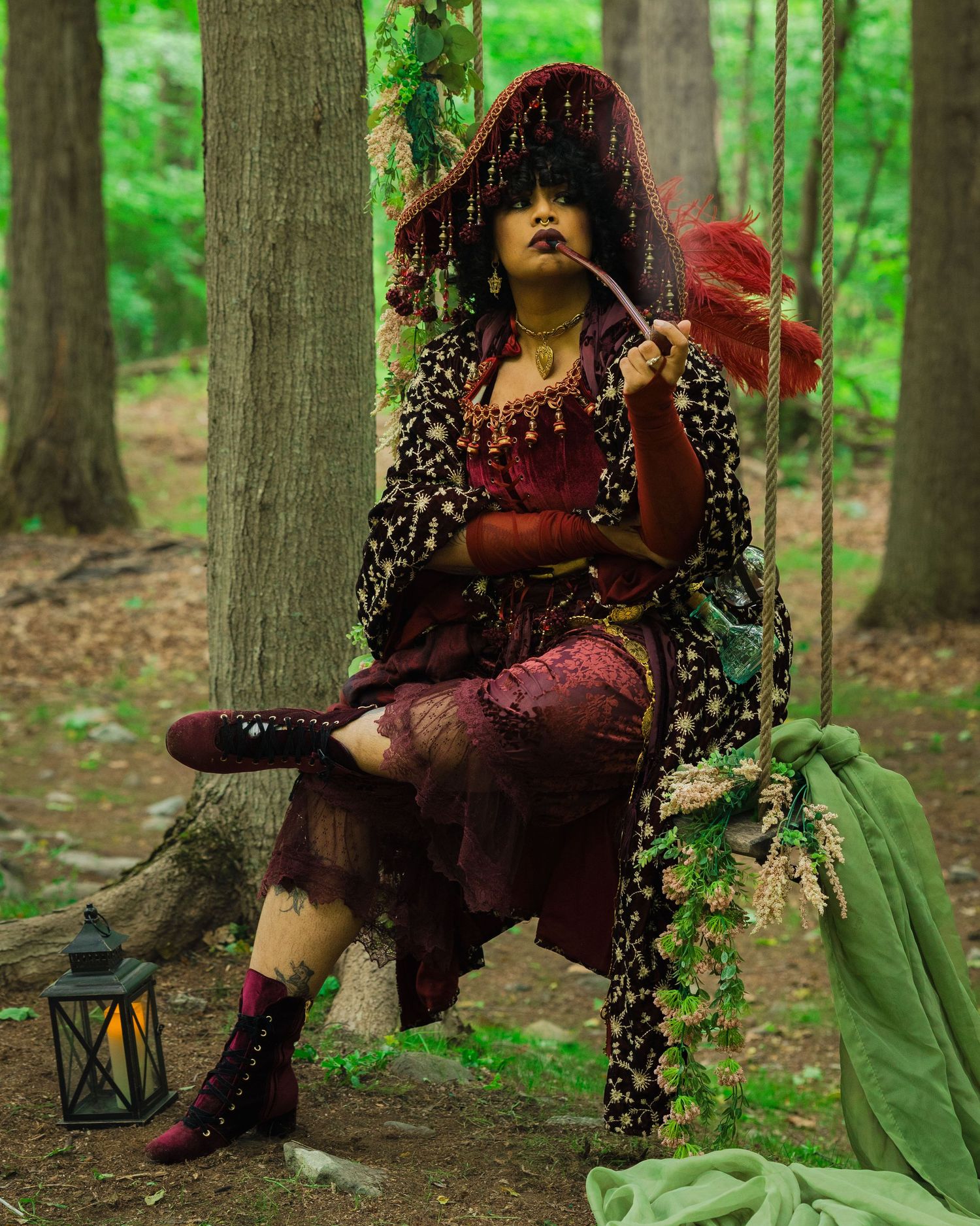 A mystical woman in ornate red and burgundy costume with veil sits on a forest swing, holding a feather fan.