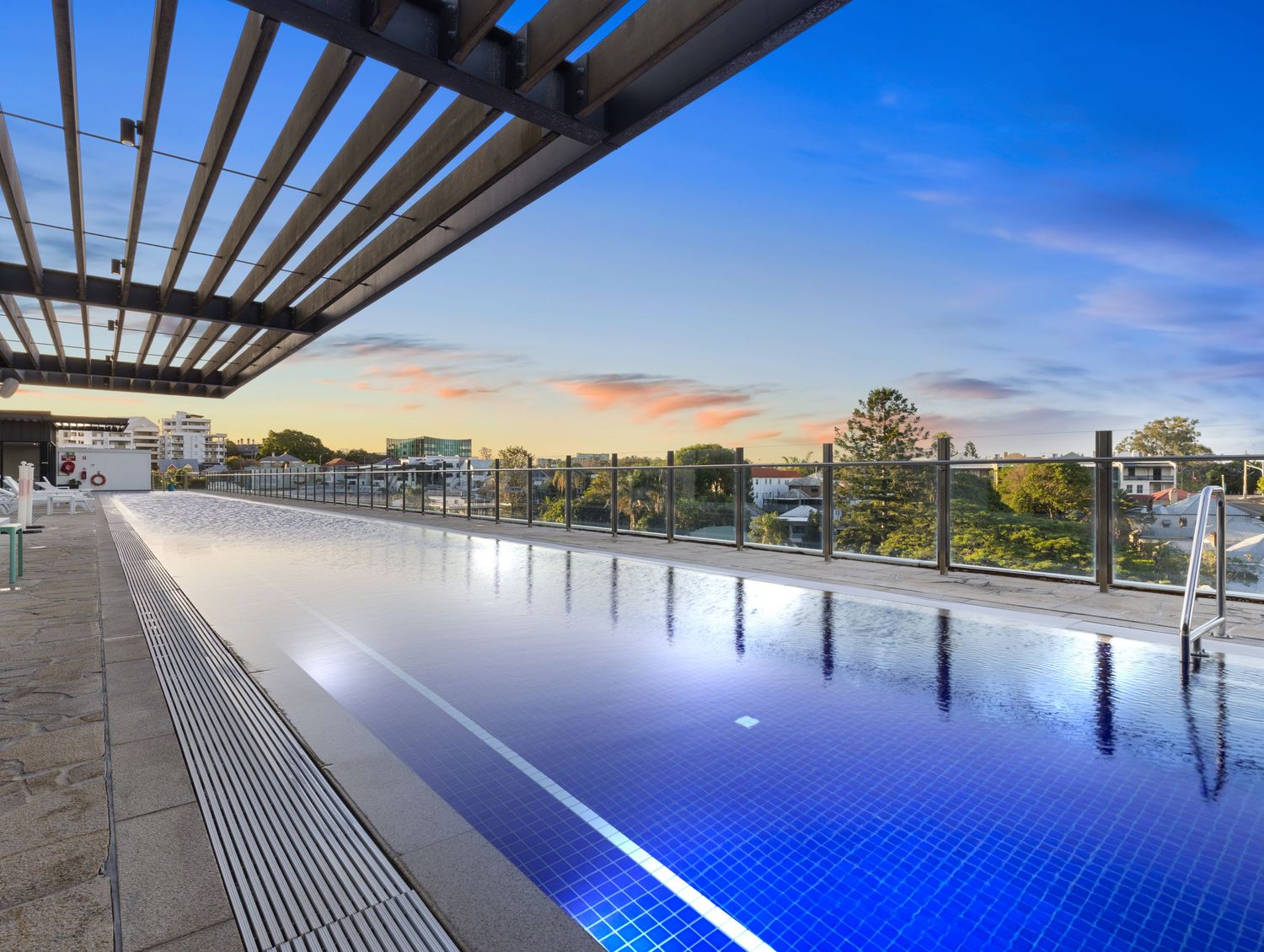Luxury infinity pool with blue lighting and pergola overlooking city views at sunset.