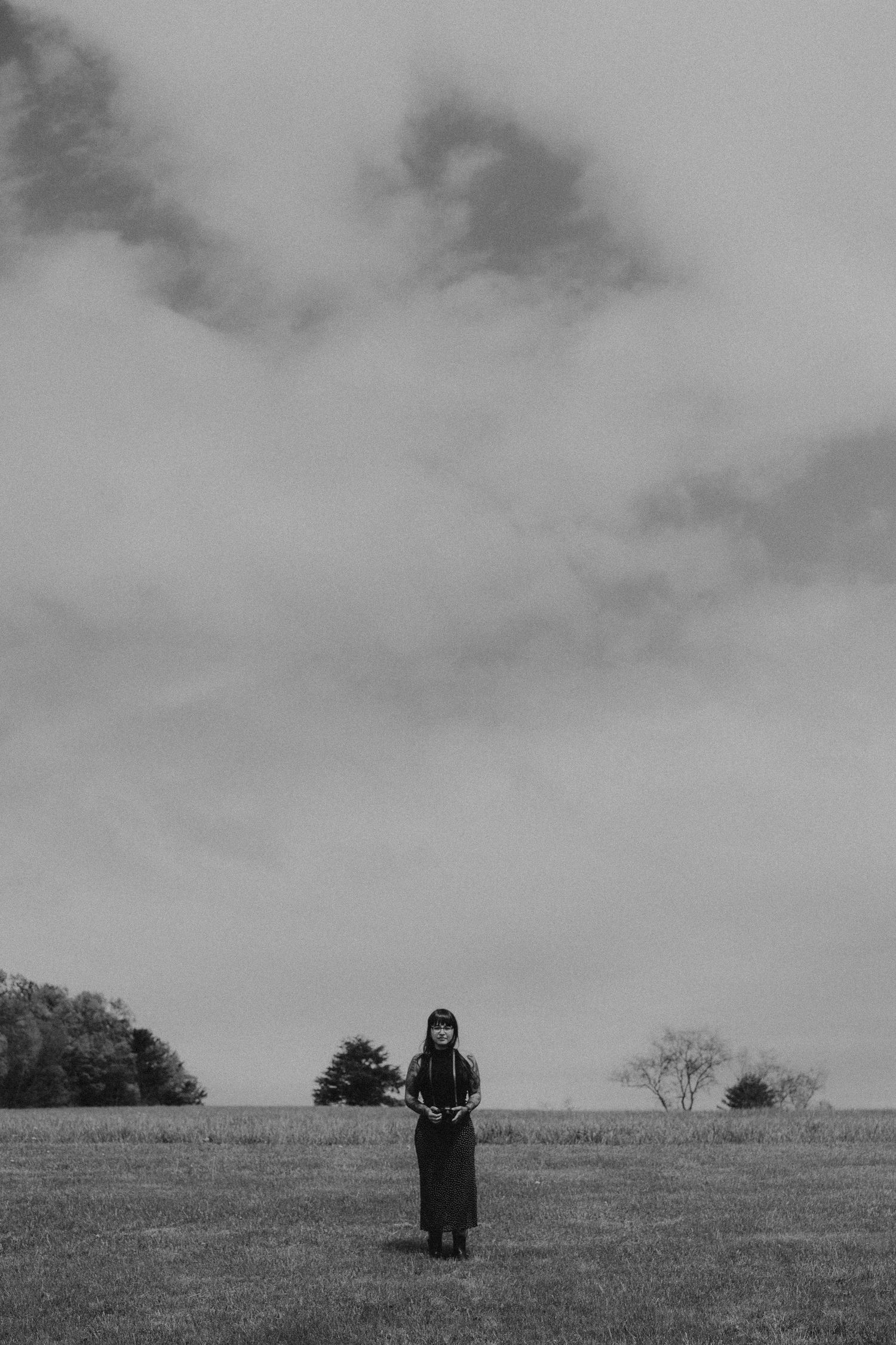 A figure stands in a dark coat on a grassy hillside against a moody overcast sky in black and white.