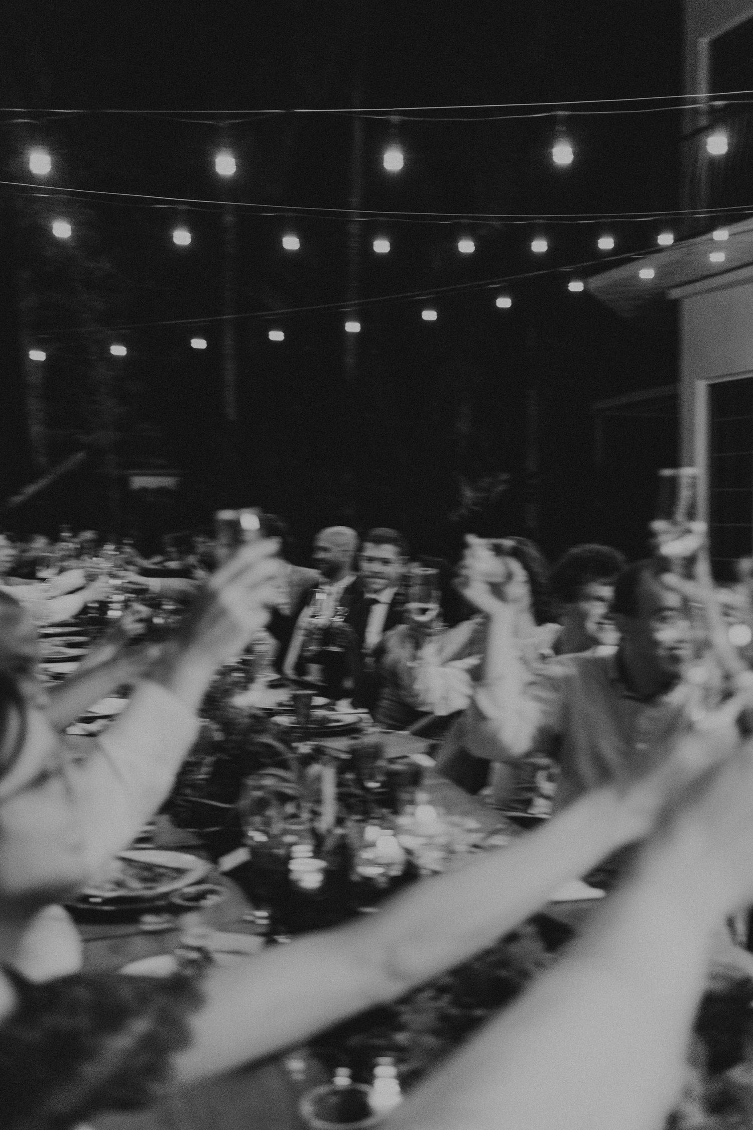 String lights illuminate an evening wedding reception with raised hands in black and white.