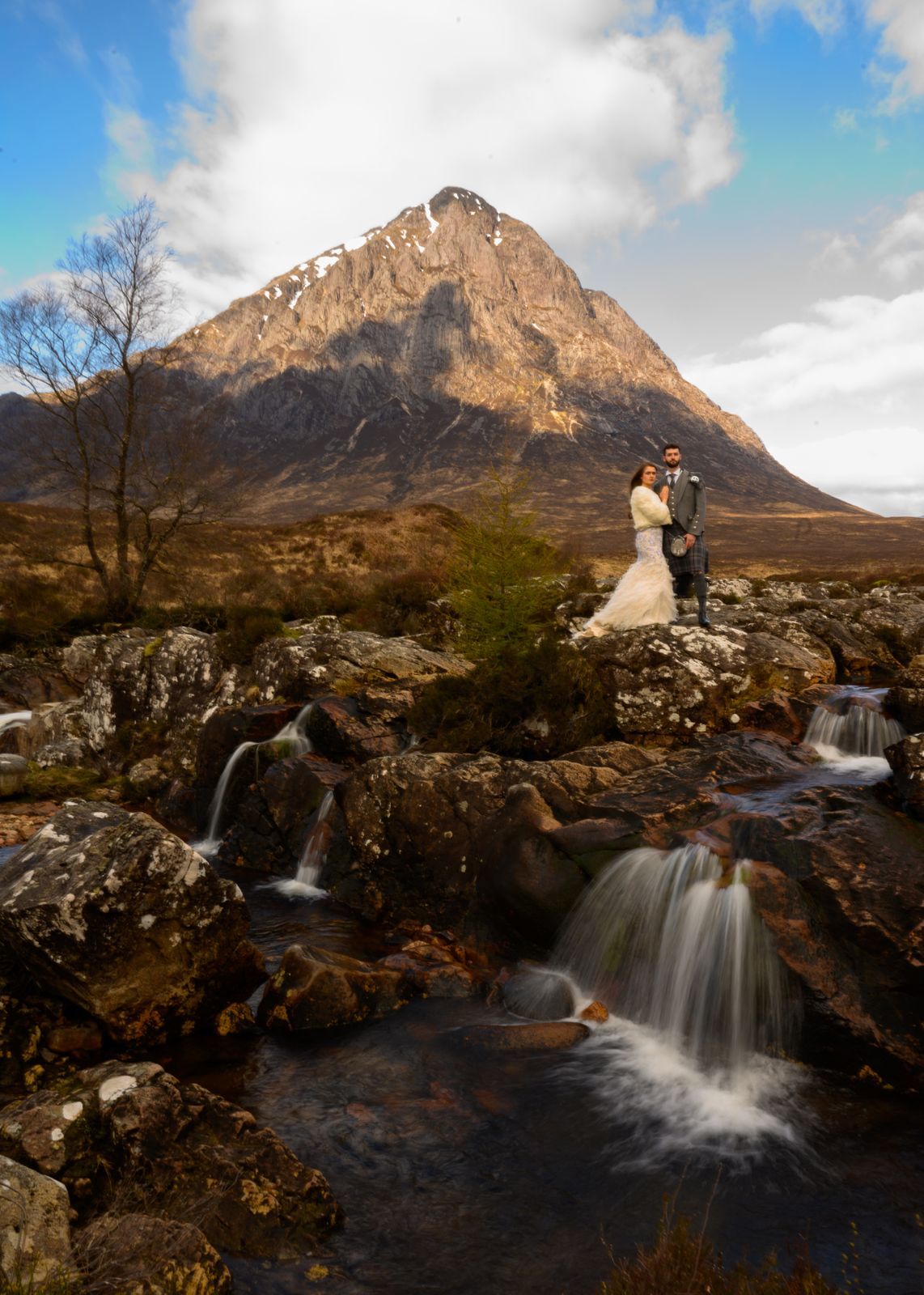 Couple embracing on rocks by a waterfall with Buachaille Etive Mor mountain in the Scottish Highlands.